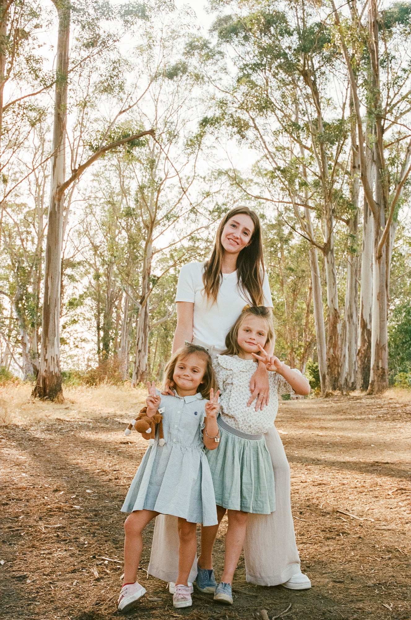 Mother stands smiling between two young daughters in matching light dresses on a sun-dappled forest path lined with tall eucalyptus trees