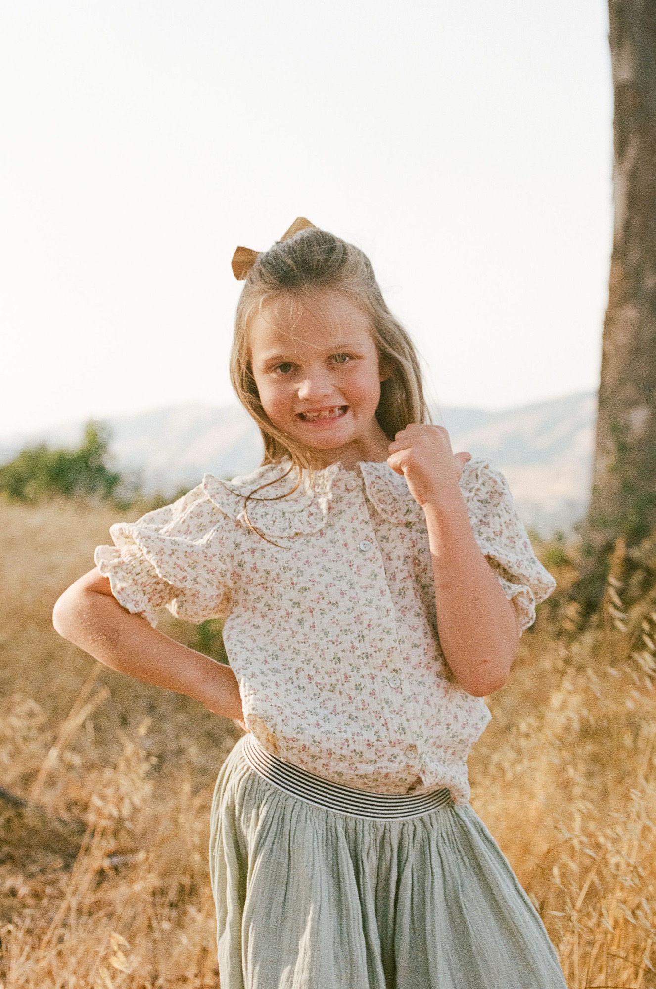 A young girl with a cheerful smile stands in golden grassland, wearing a delicate floral top with puffed sleeves and a soft sage skirt