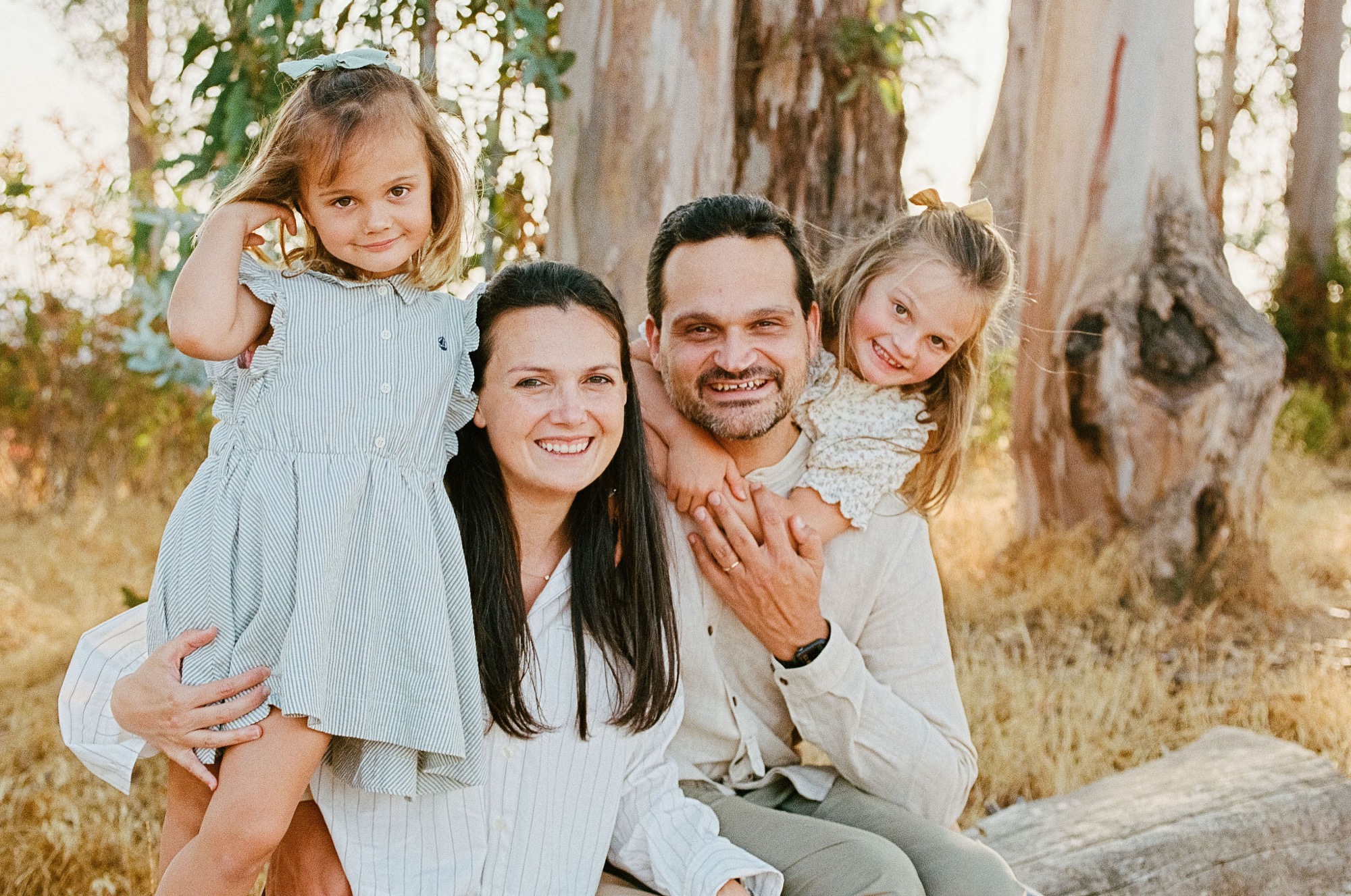 A smiling family of four poses together among weathered tree trunks in golden afternoon light, with two young daughters perched on their parents' shoulders
