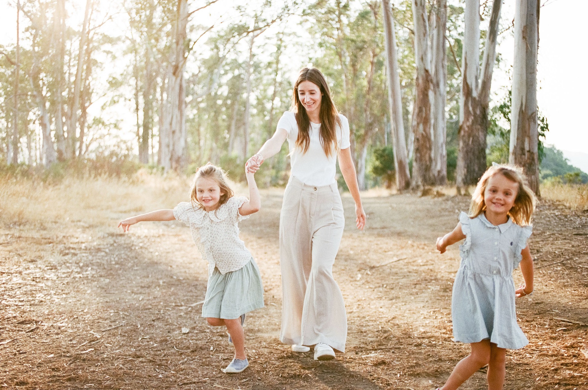 A woman walks hand-in-hand with two young girls along a sun-dappled dirt path beneath towering eucalyptus trees, all three smiling and moving together