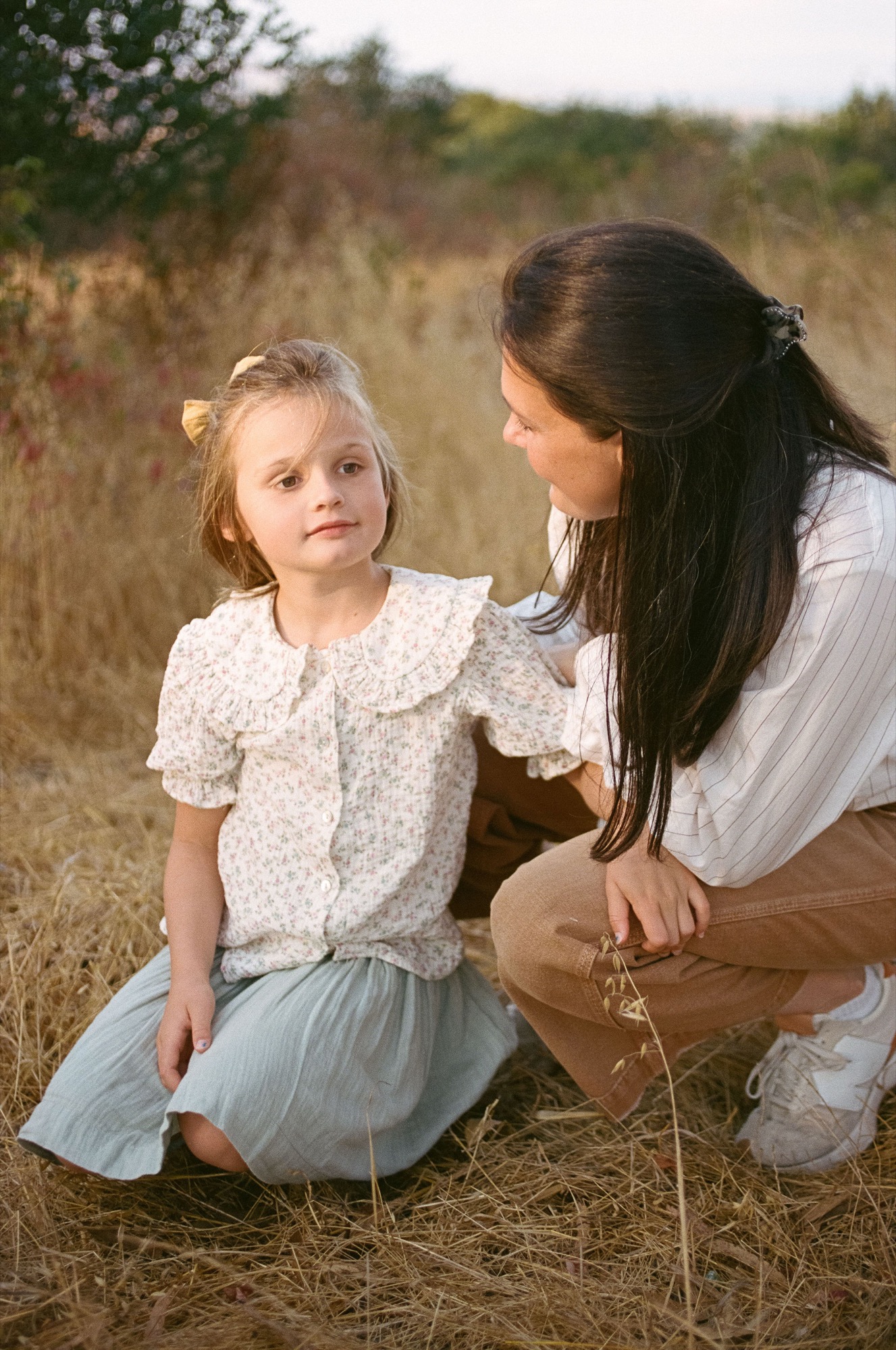 A woman crouches beside a young girl in a sunlit meadow, gently touching her shoulder while the child gazes thoughtfully away