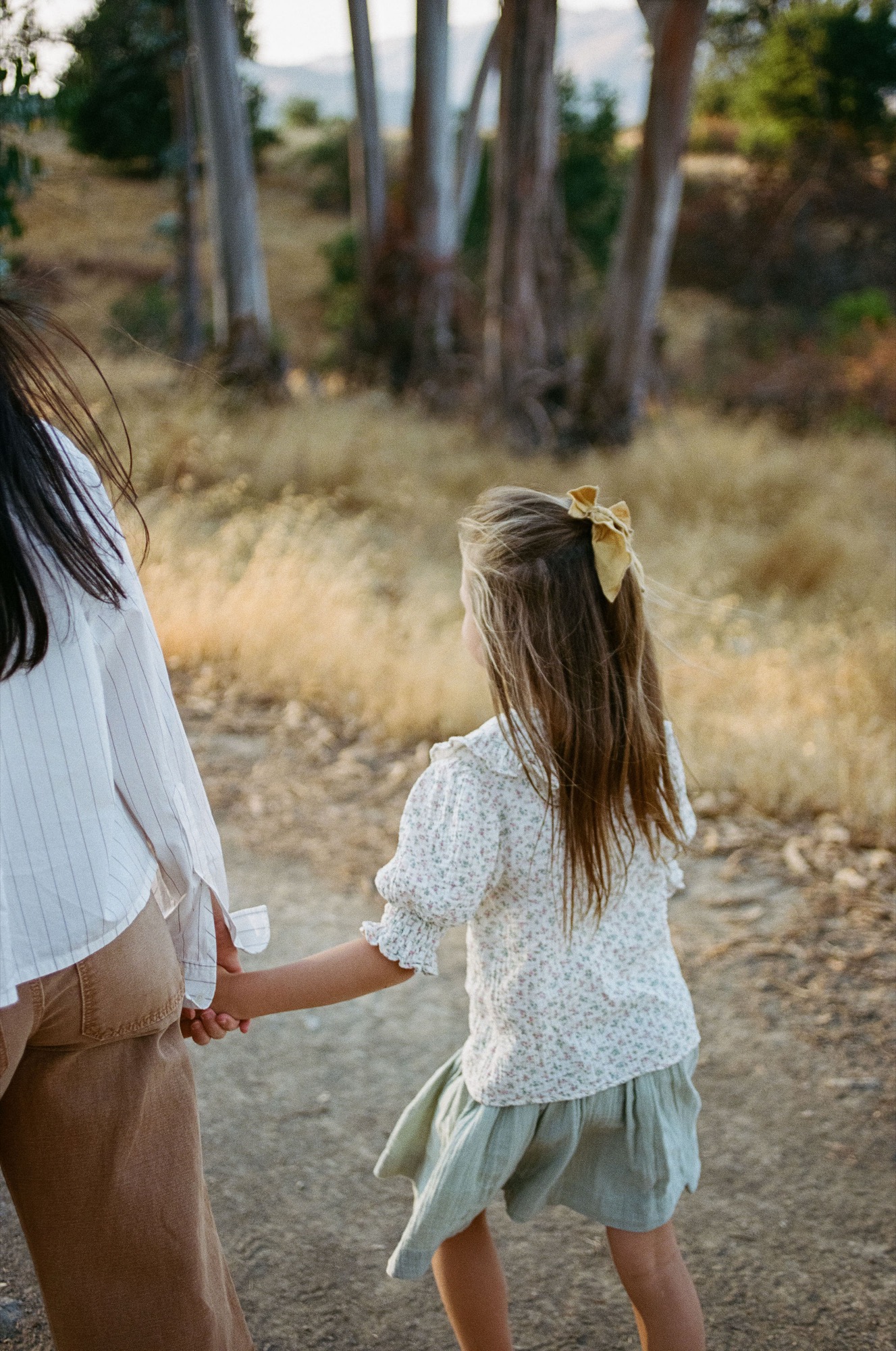 An adult and child walk hand-in-hand down a dusty trail, the child's blonde hair catching afternoon light as dry grass and eucalyptus trees blur behind them