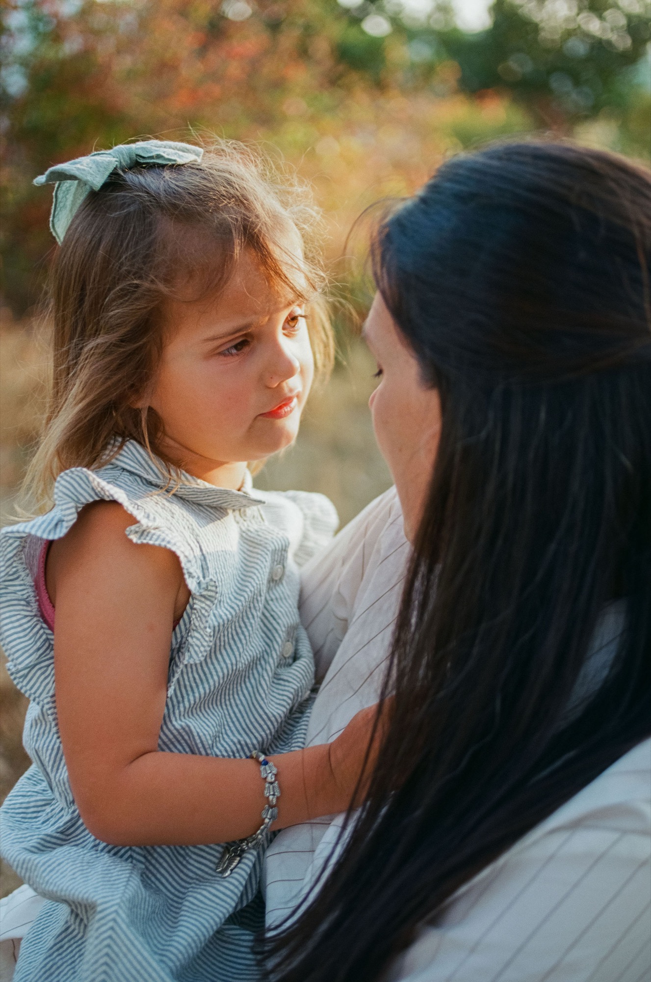 A woman holds her young daughter close, both gazing at each other with warm affection as golden afternoon light filters through the trees