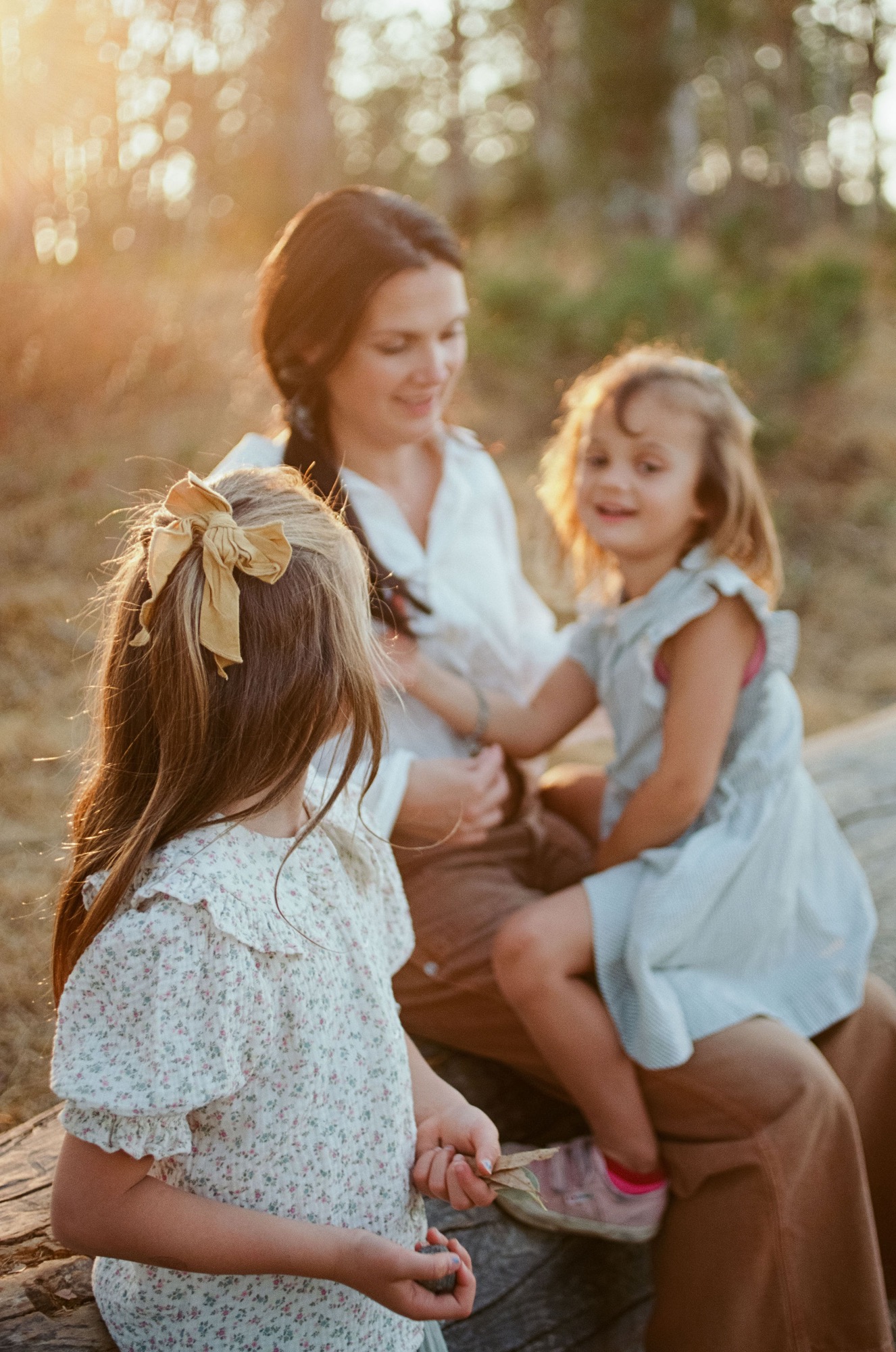 A mother kneels among golden sunlight, holding two daughters close on a wooded trail, their expressions warm and connected
