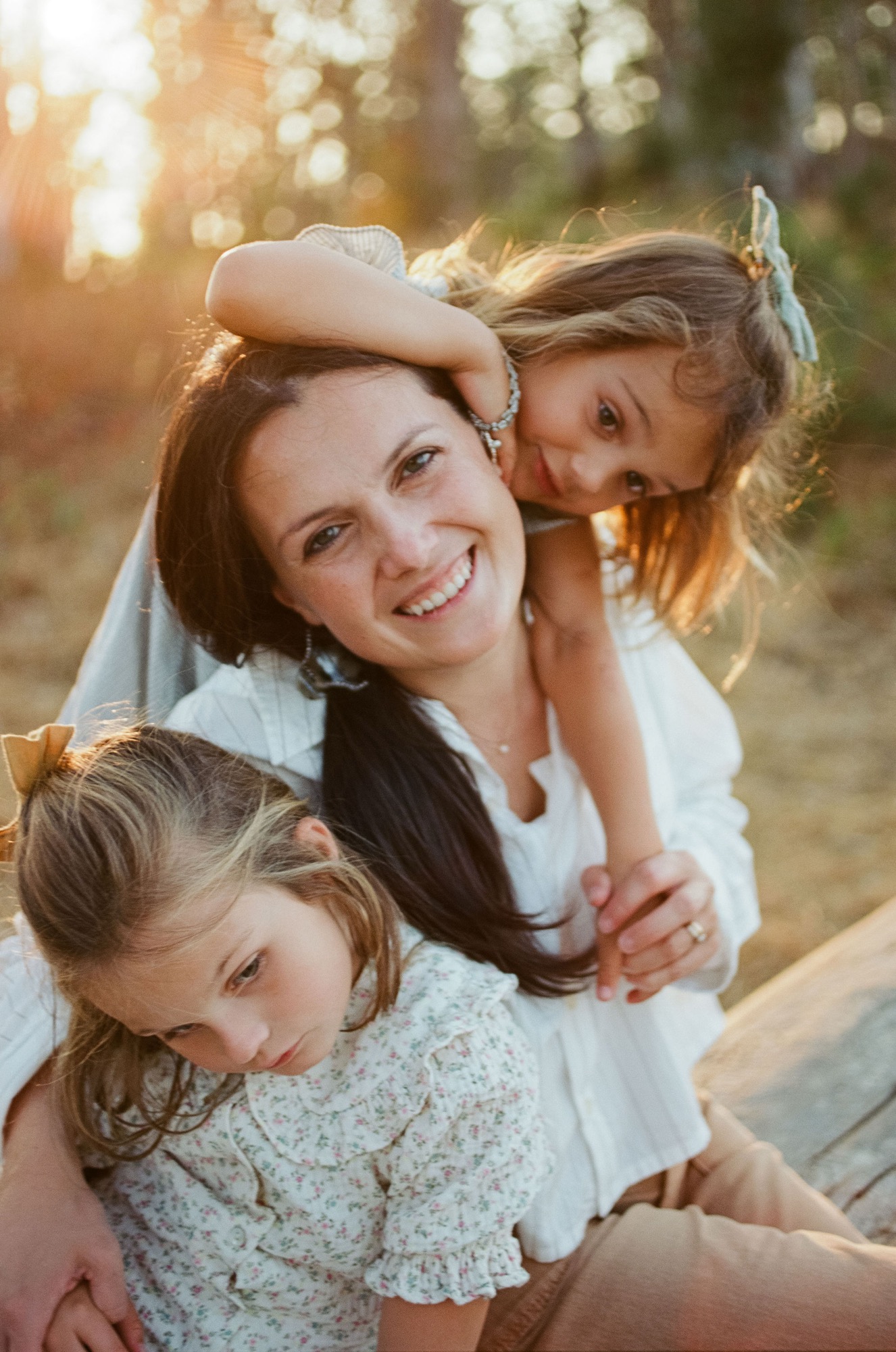 Mother hugs two young daughters from behind in golden afternoon light, all three smiling warmly at the camera