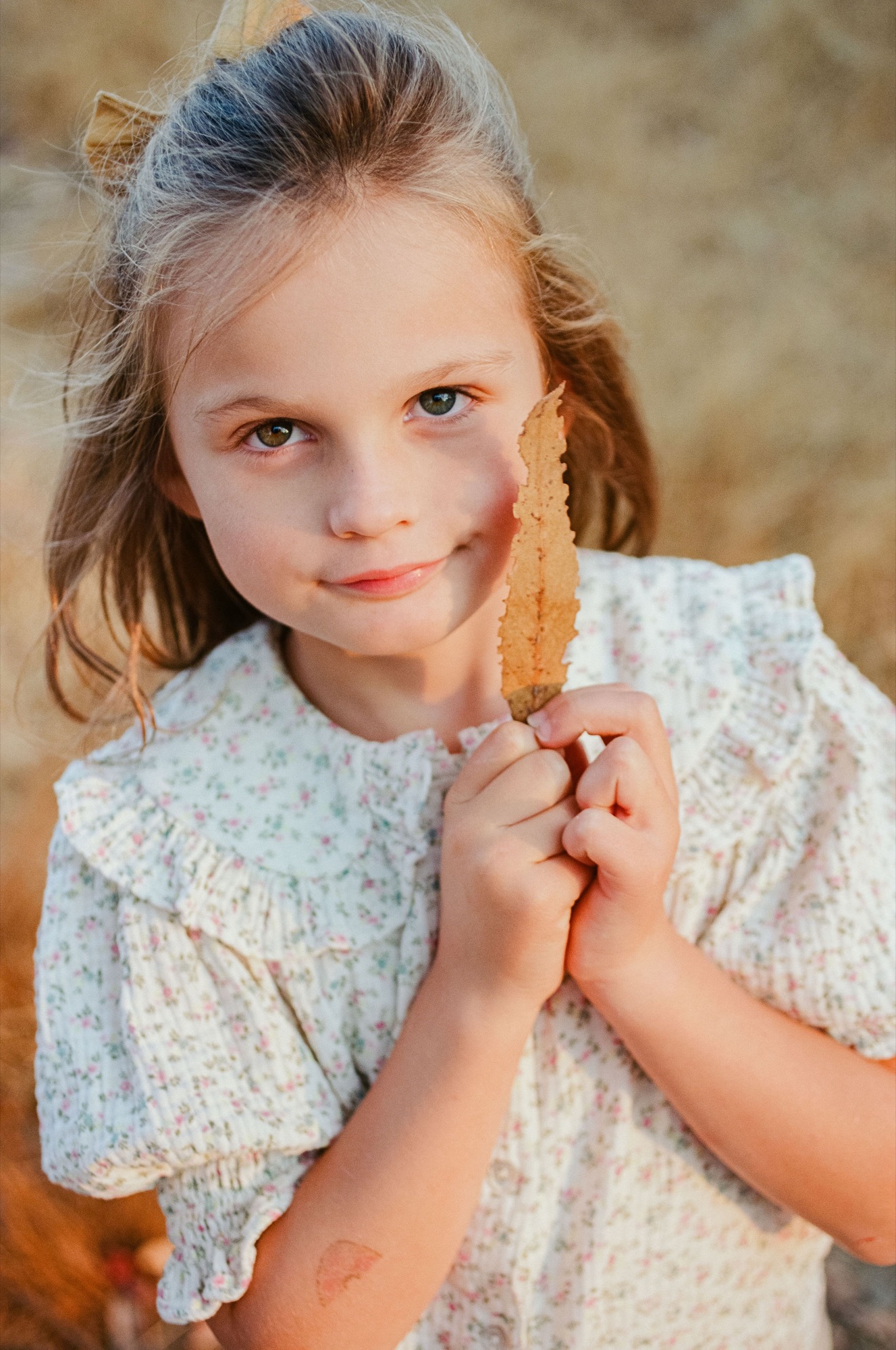 Young girl in a light patterned dress holds a golden-brown leaf, gazing directly at the camera with a gentle expression against a soft, blurred natural background