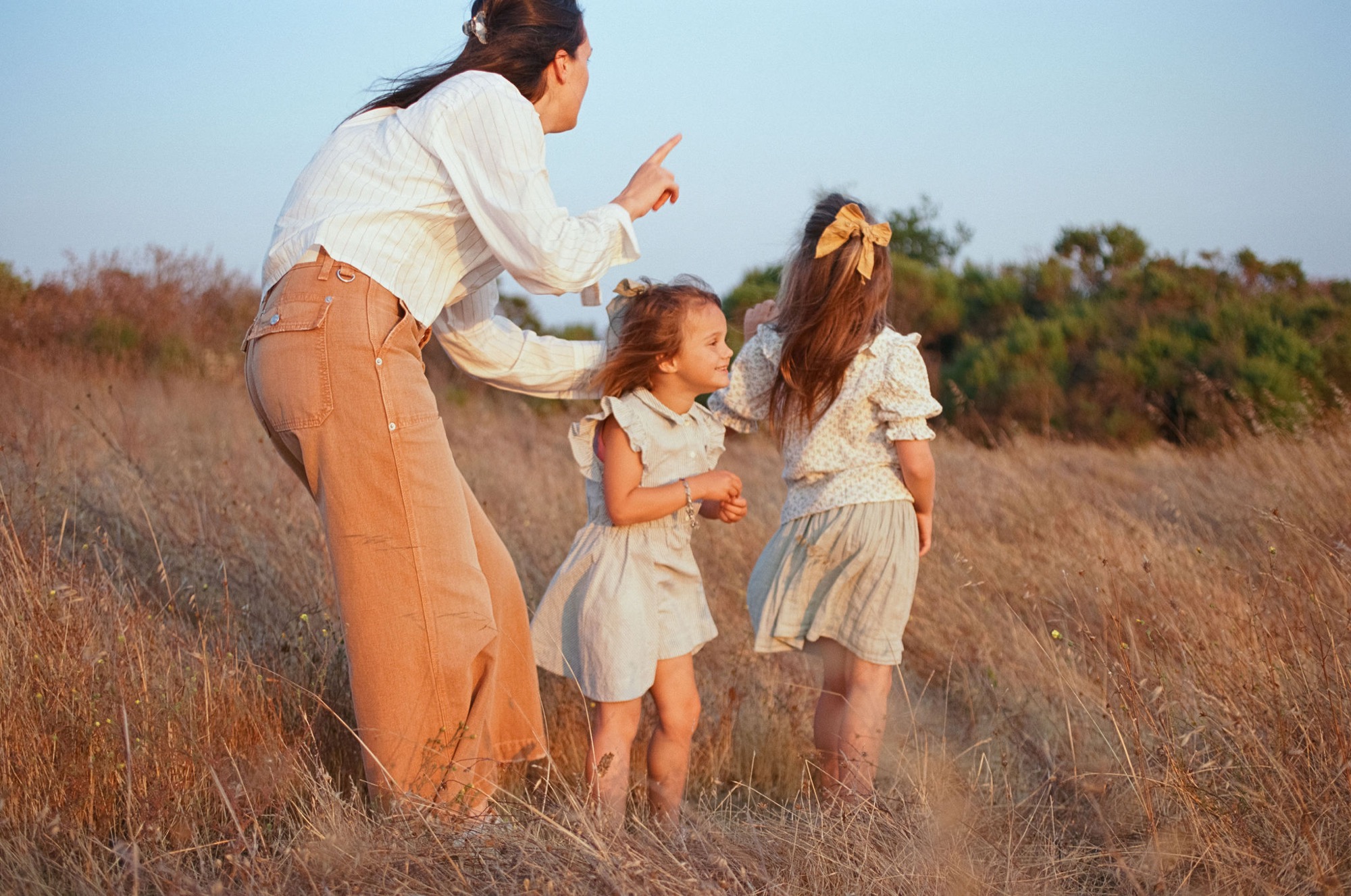 A woman plays with two young children in golden dry grass, their silhouettes backlit by warm afternoon sunlight across an open meadow