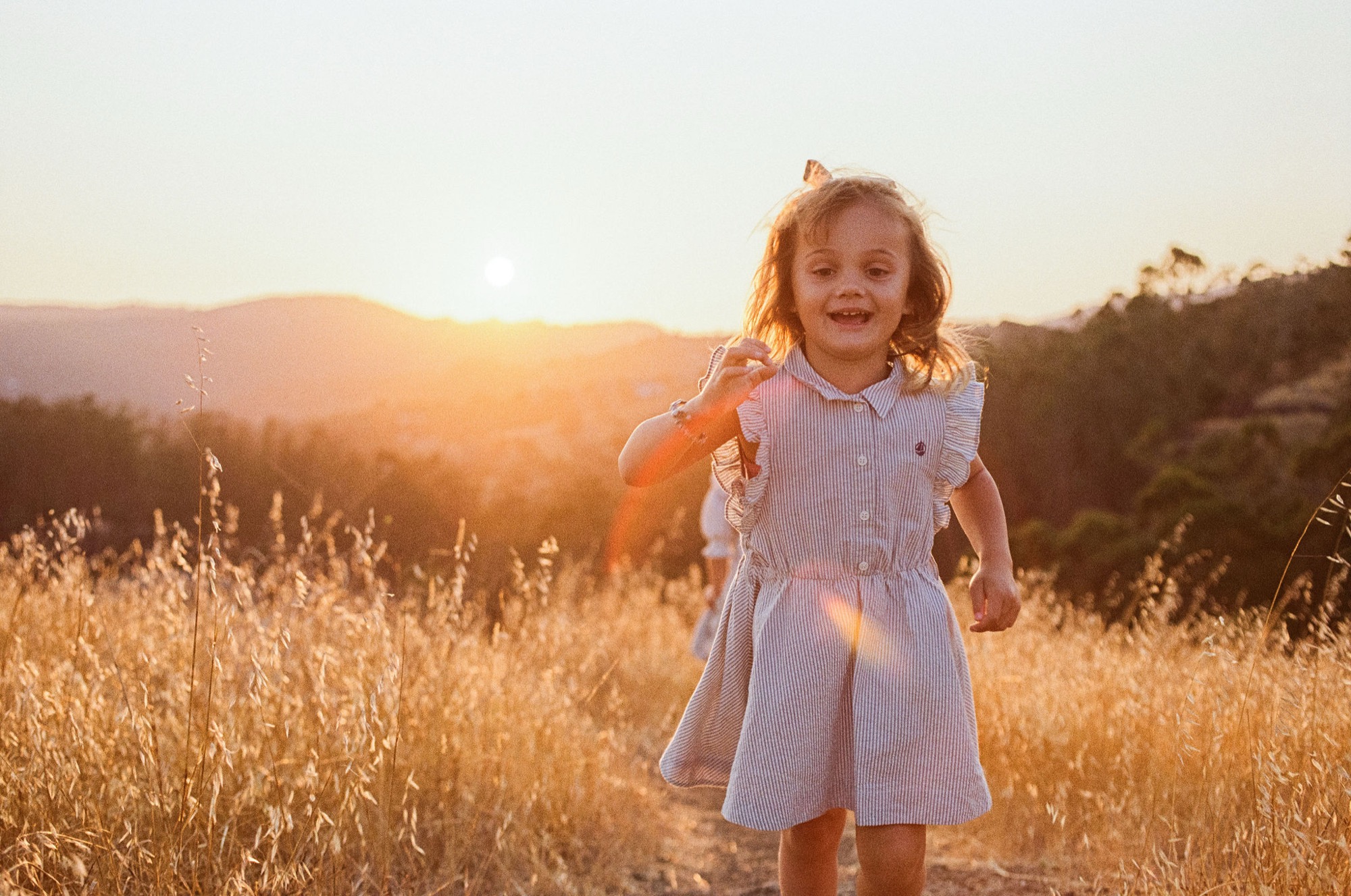 A young child with golden hair runs through a sunlit golden field at sunset, arms outstretched with genuine joy and abandon