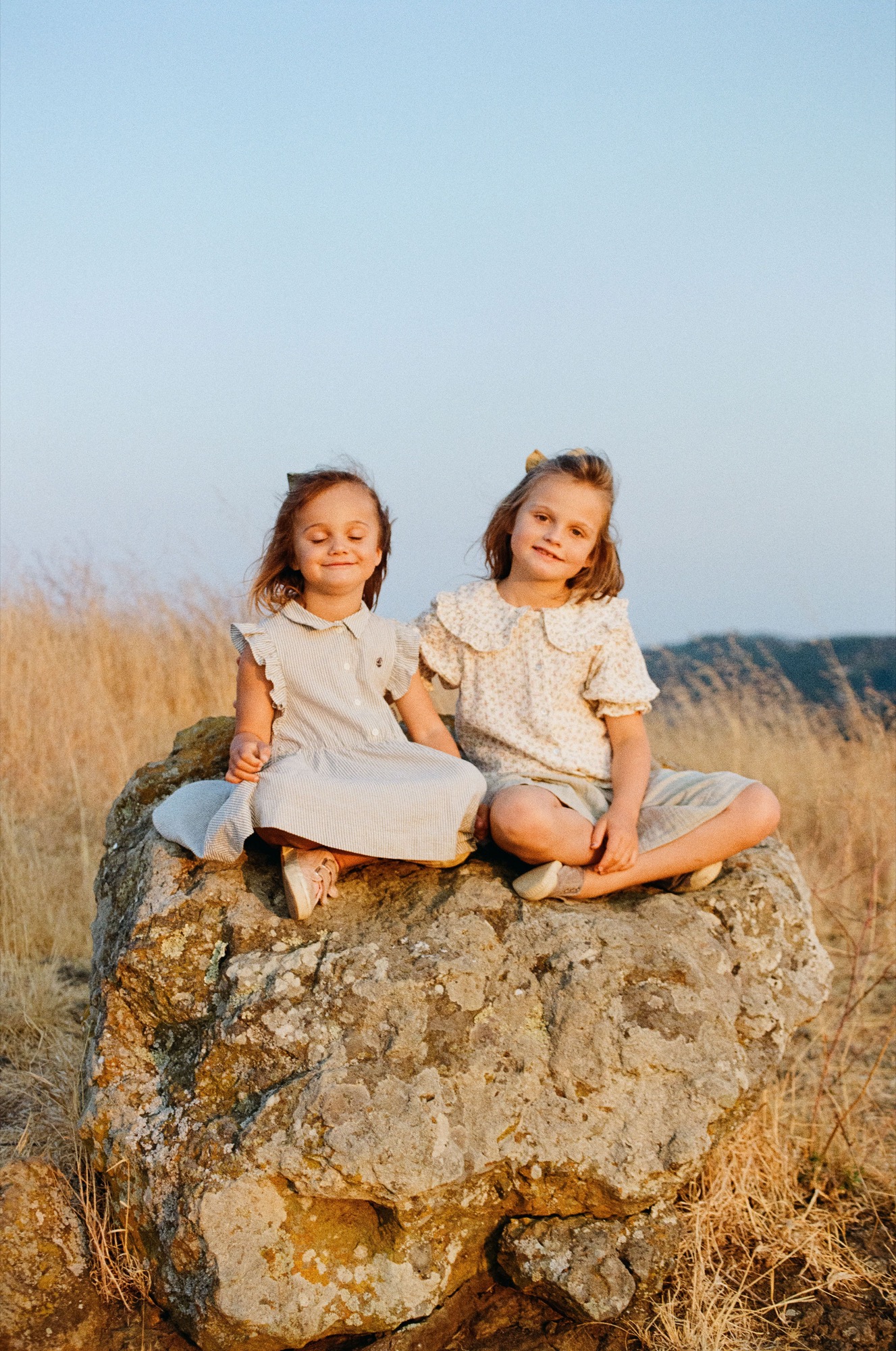 Two young girls sit barefoot atop a large weathered boulder, squinting slightly in golden afternoon light with dry hillside grasses stretching behind them