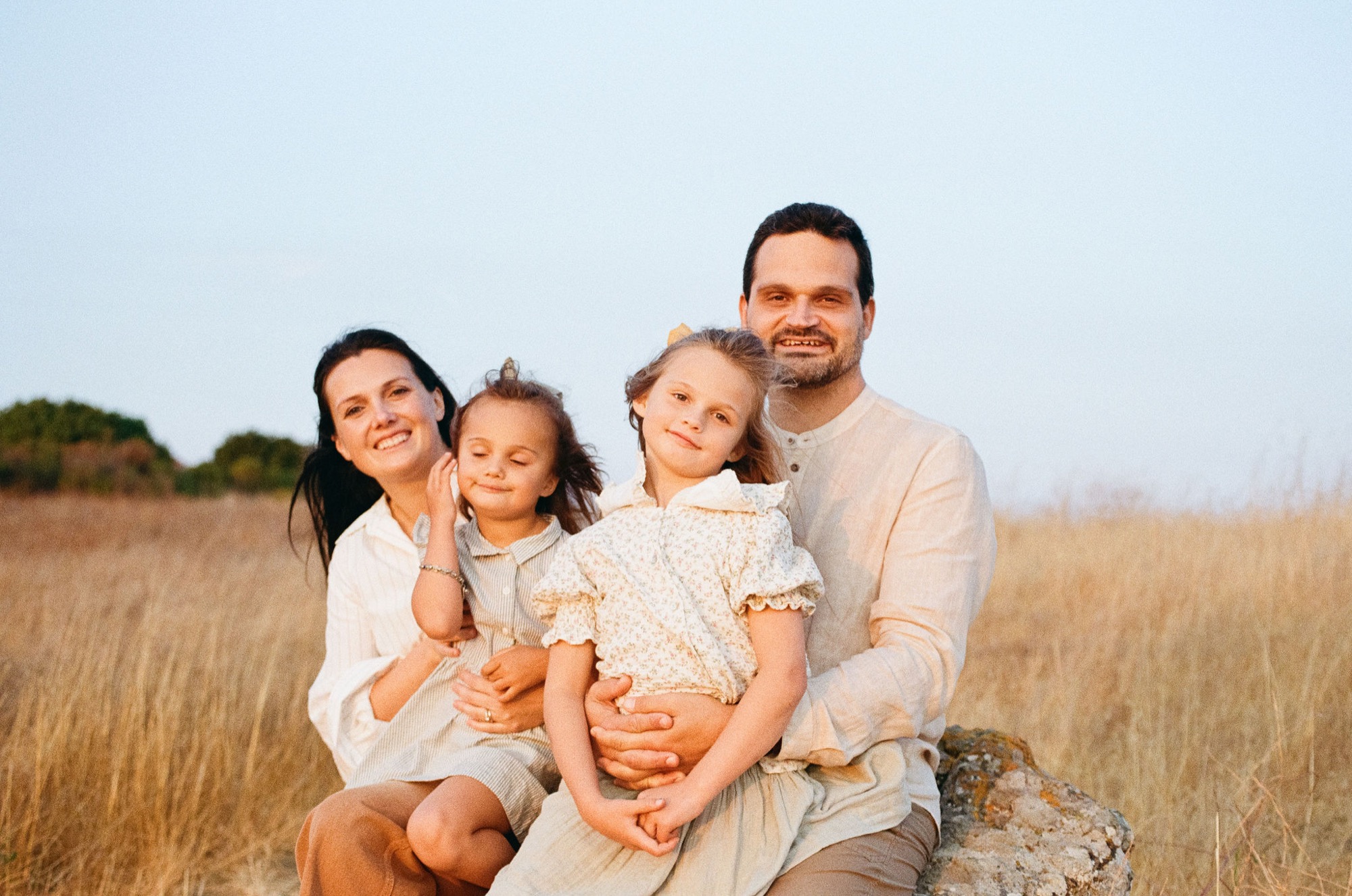 Family of four seated together in golden meadow grass, smiling warmly at camera with soft afternoon light illuminating their peaceful outdoor moment