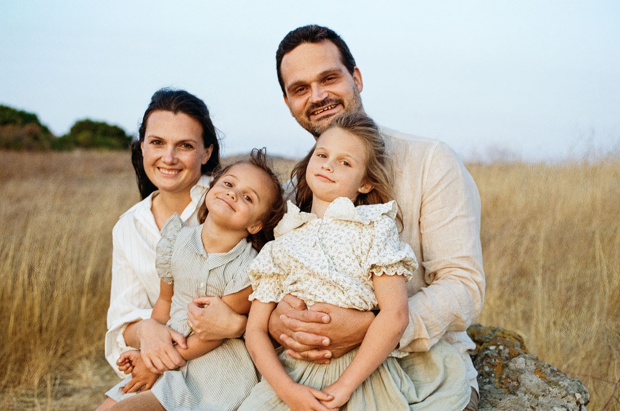 A family of four smiles warmly in a golden field, with the father and mother holding their two children close as evening light bathes the open landscape