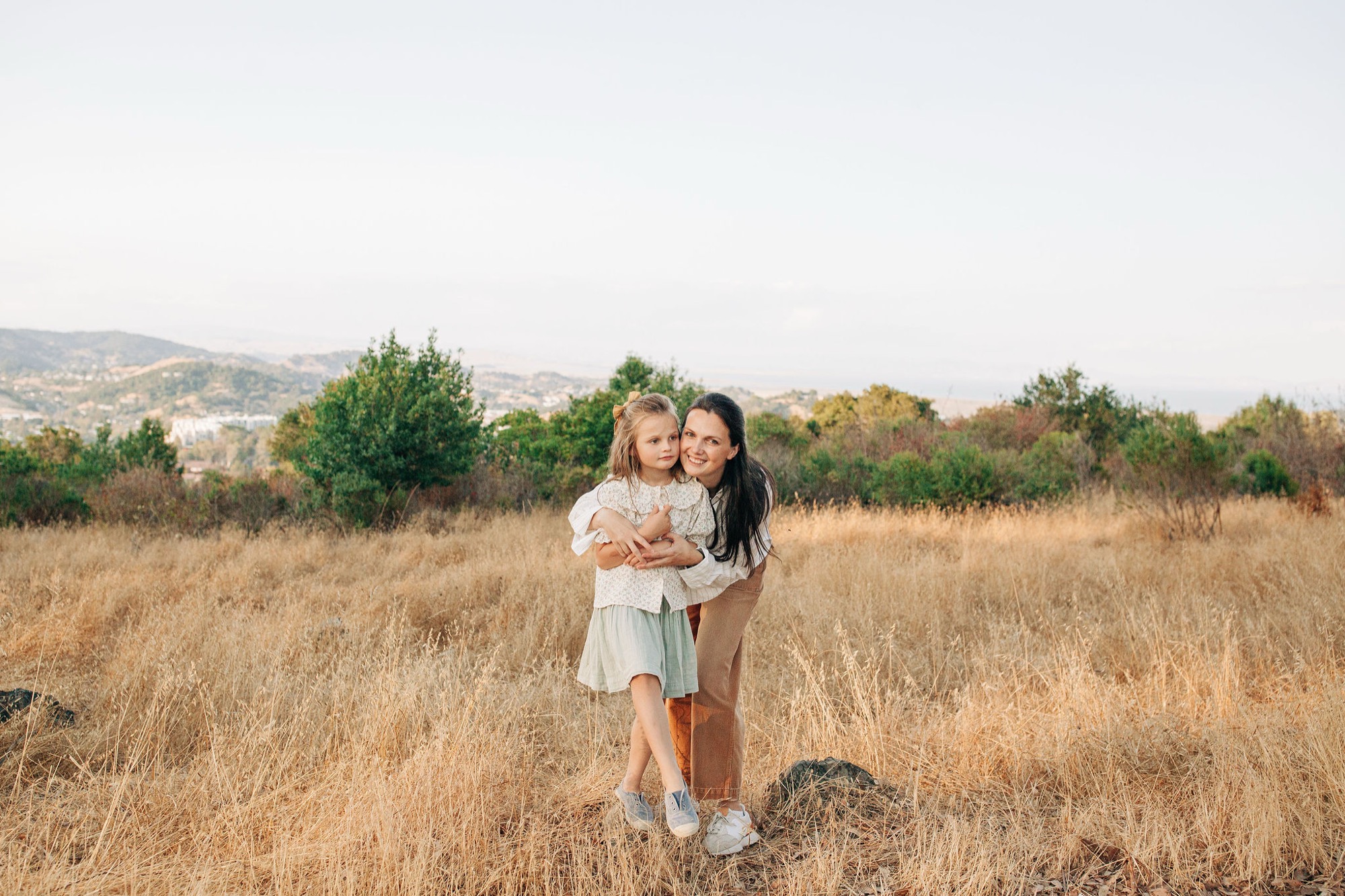 Two young girls embrace warmly in golden grassland, their genuine smiles and relaxed postures suggesting close sisterly affection beneath soft afternoon light