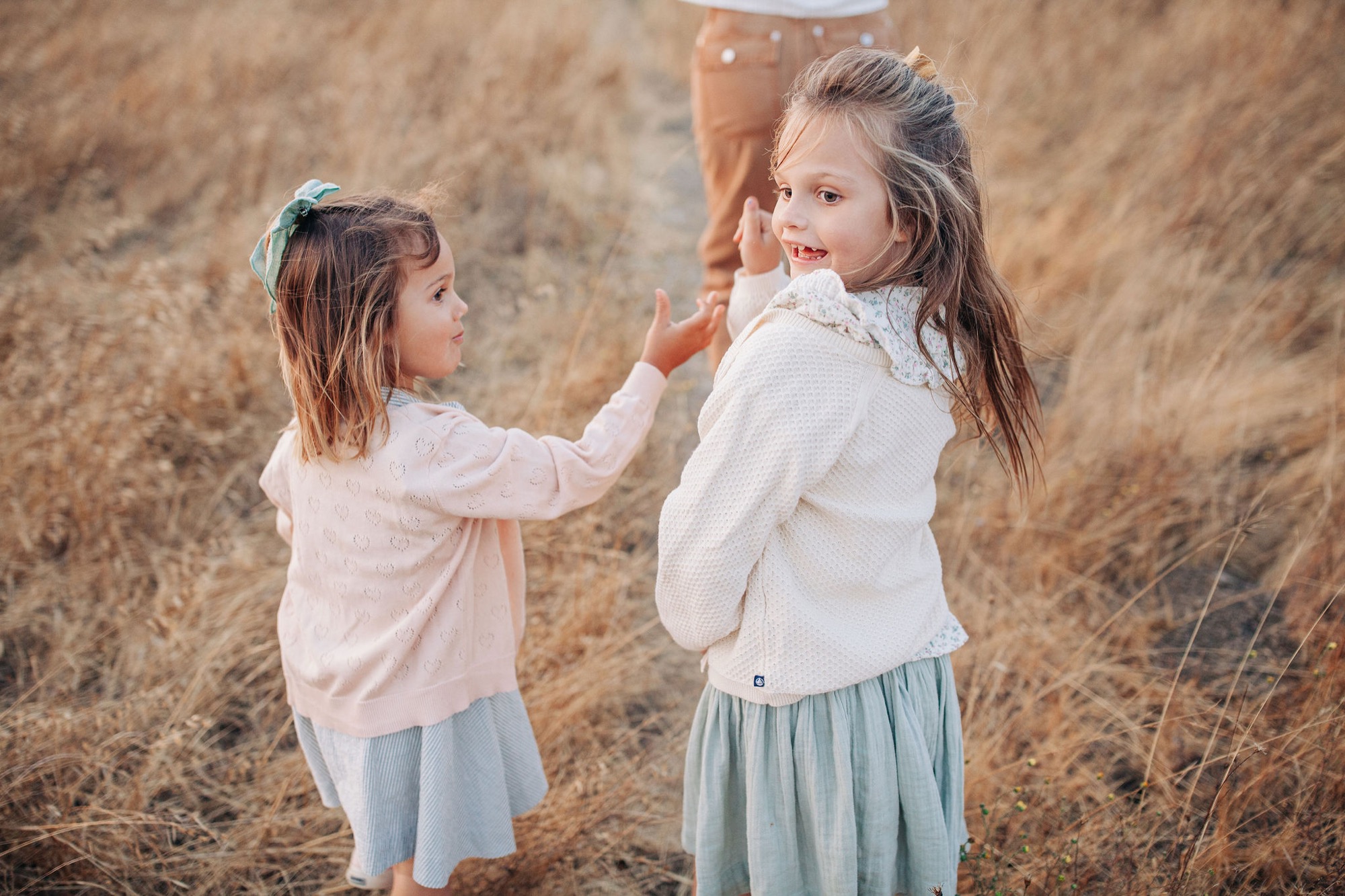 Two young girls hold hands on a dry hillside, their white tops glowing against brown grass as they turn toward the camera with bright smiles
