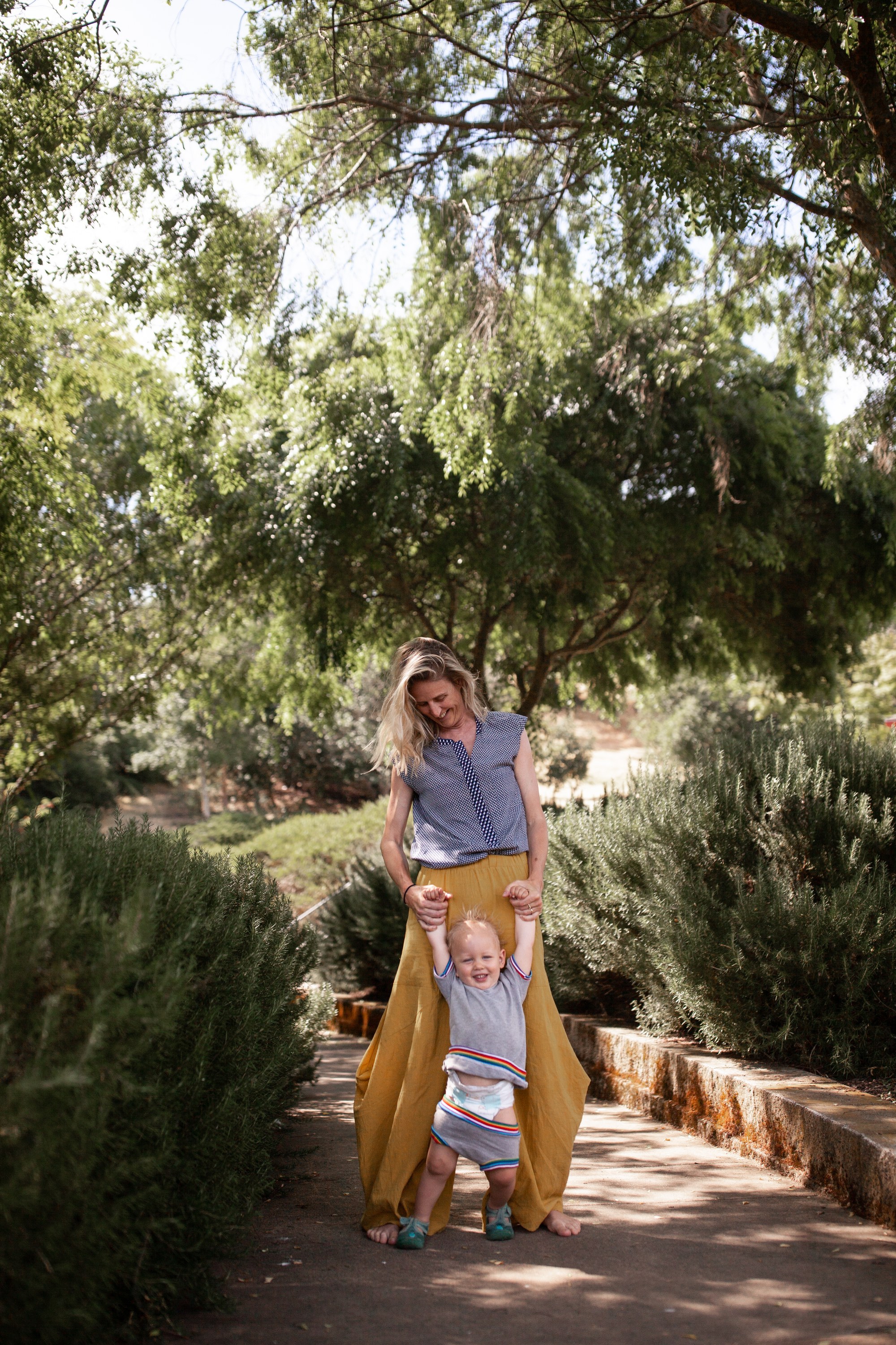 A toddler takes assisted steps down a path lined with rosemary hedges, held up by his mother, at a park in Terra Linda
