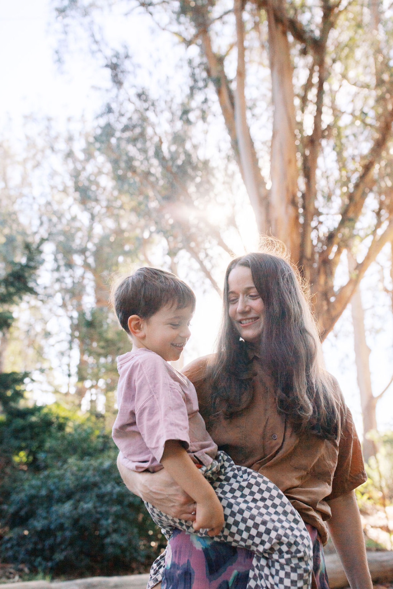 A woman in a brown shirt holds her young son against tall eucalyptus trees, their faces close together in warm afternoon sunlight filtering through the canopy