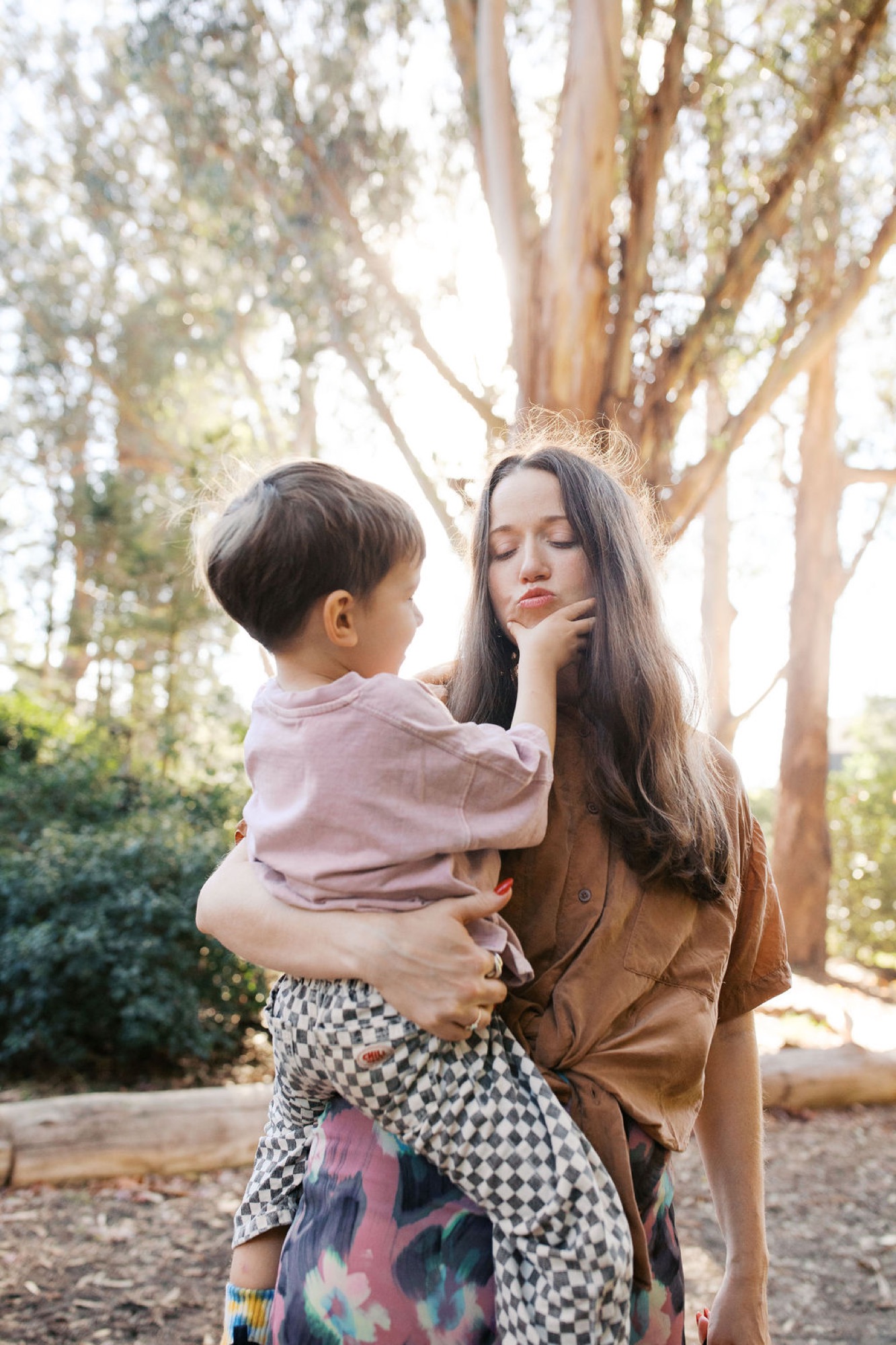 A woman with long dark hair and red lipstick holds a toddler against her chest beneath towering eucalyptus trees, bathed in warm dappled sunlight