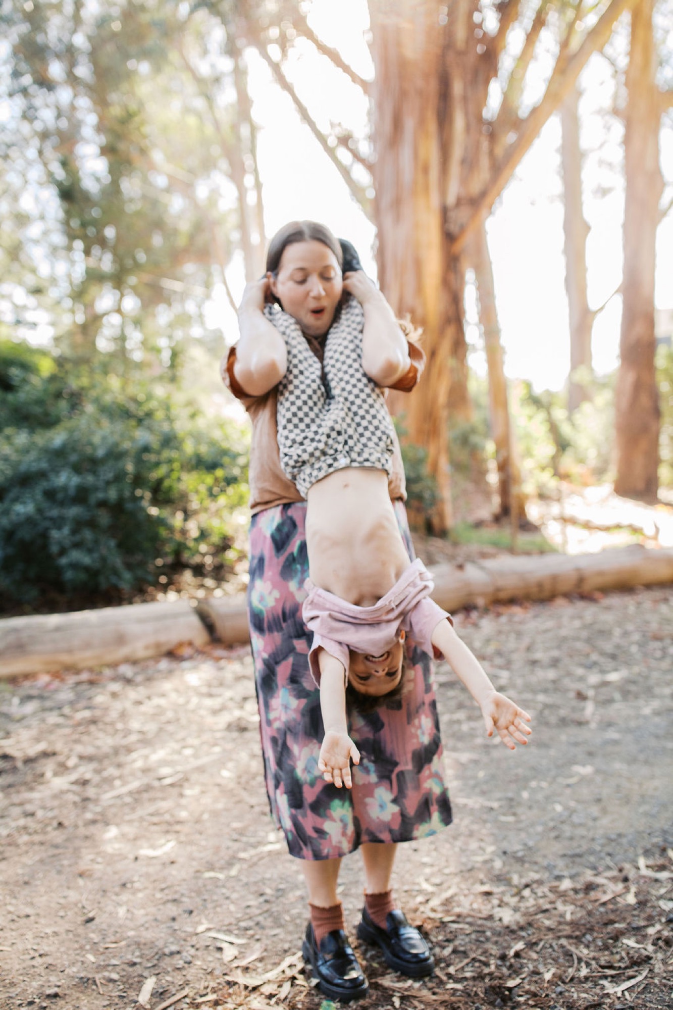 A child does a handstand on a shaded forest trail, wearing a patterned skirt and checkered shirt, with tall eucalyptus trees creating dappled light behind
