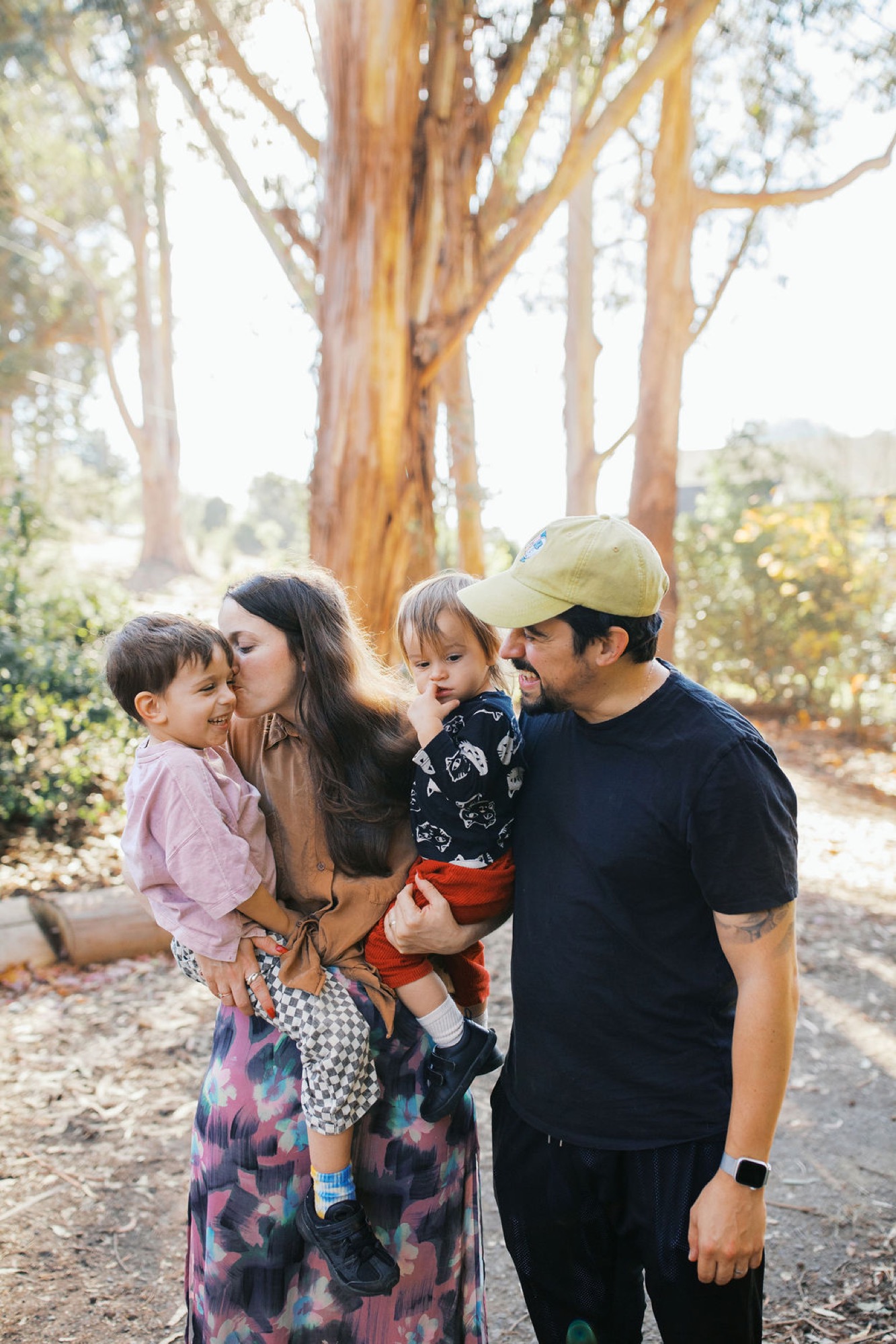 A family of four stands together among towering eucalyptus trees, with warm dappled light filtering through the canopy onto their relaxed poses