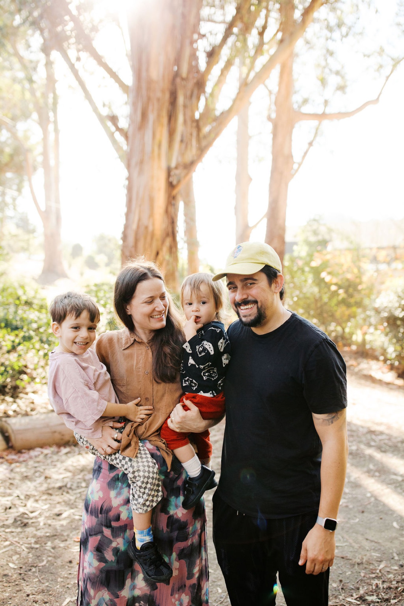 A family of four stands together on a shaded trail, bathed in golden sunlight filtering through tall eucalyptus trees, all smiling warmly at the camera