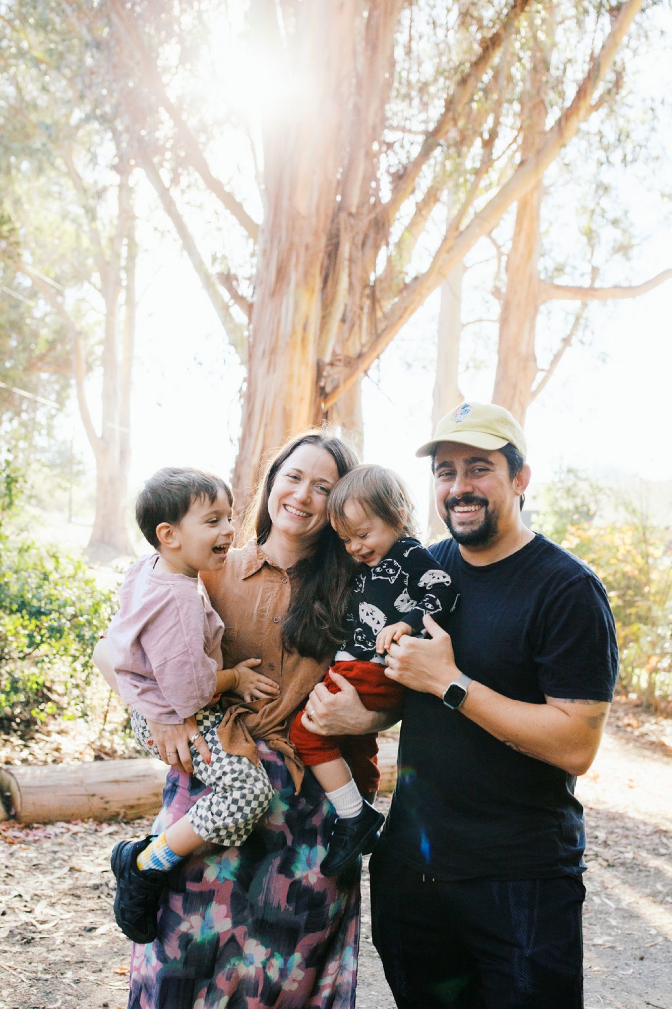 Family of four laughs together beneath towering eucalyptus trees, dappled sunlight filtering through branches onto the shaded trail
