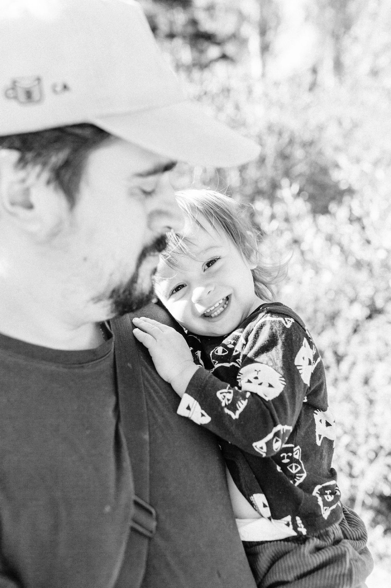 A beaming child nestled against an adult's shoulder, both bathed in dappled sunlight filtering through blooming branches