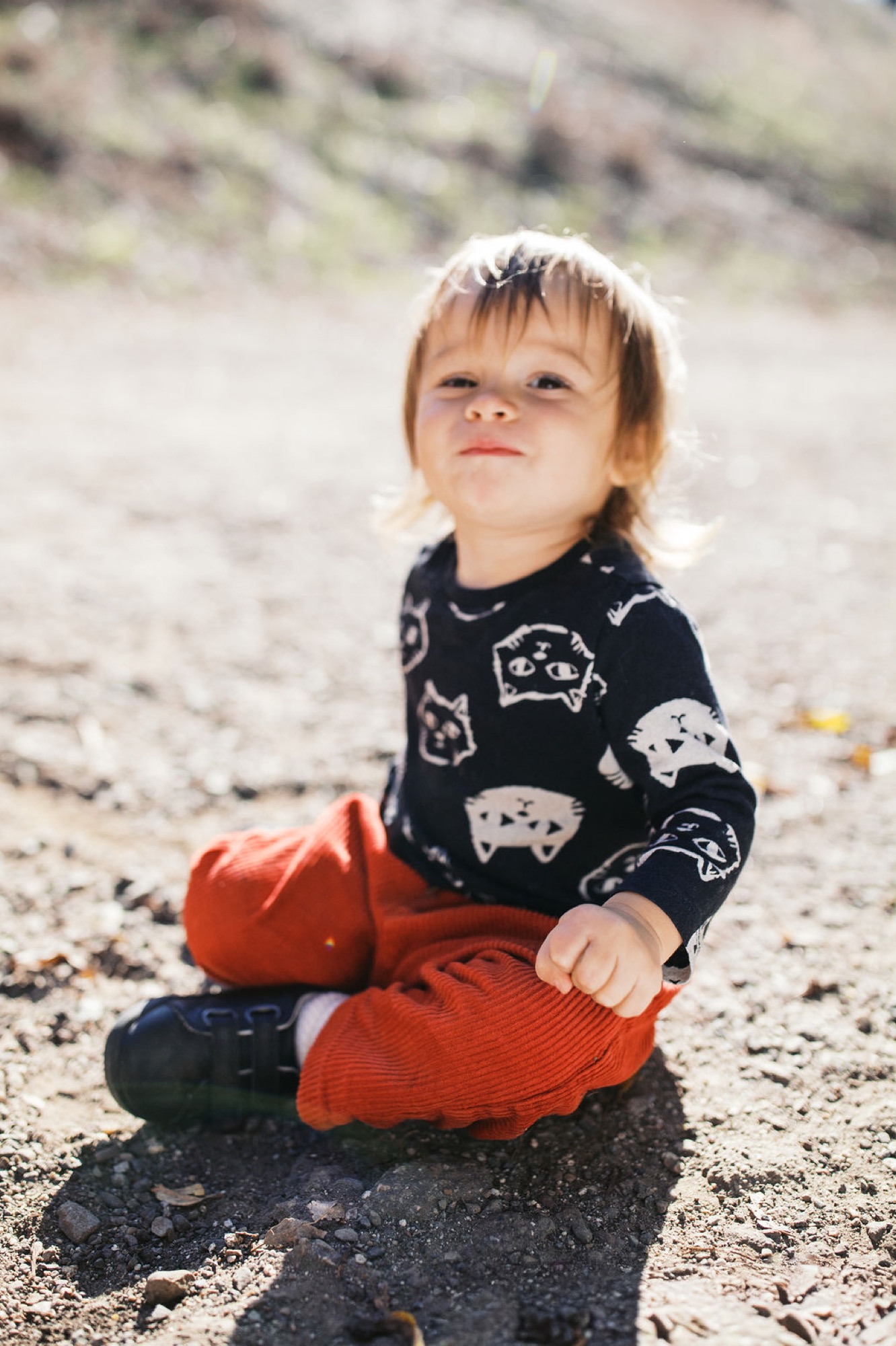 Blonde toddler in patterned black sweater sits on rocky ground with serious expression, golden afternoon light illuminating their face