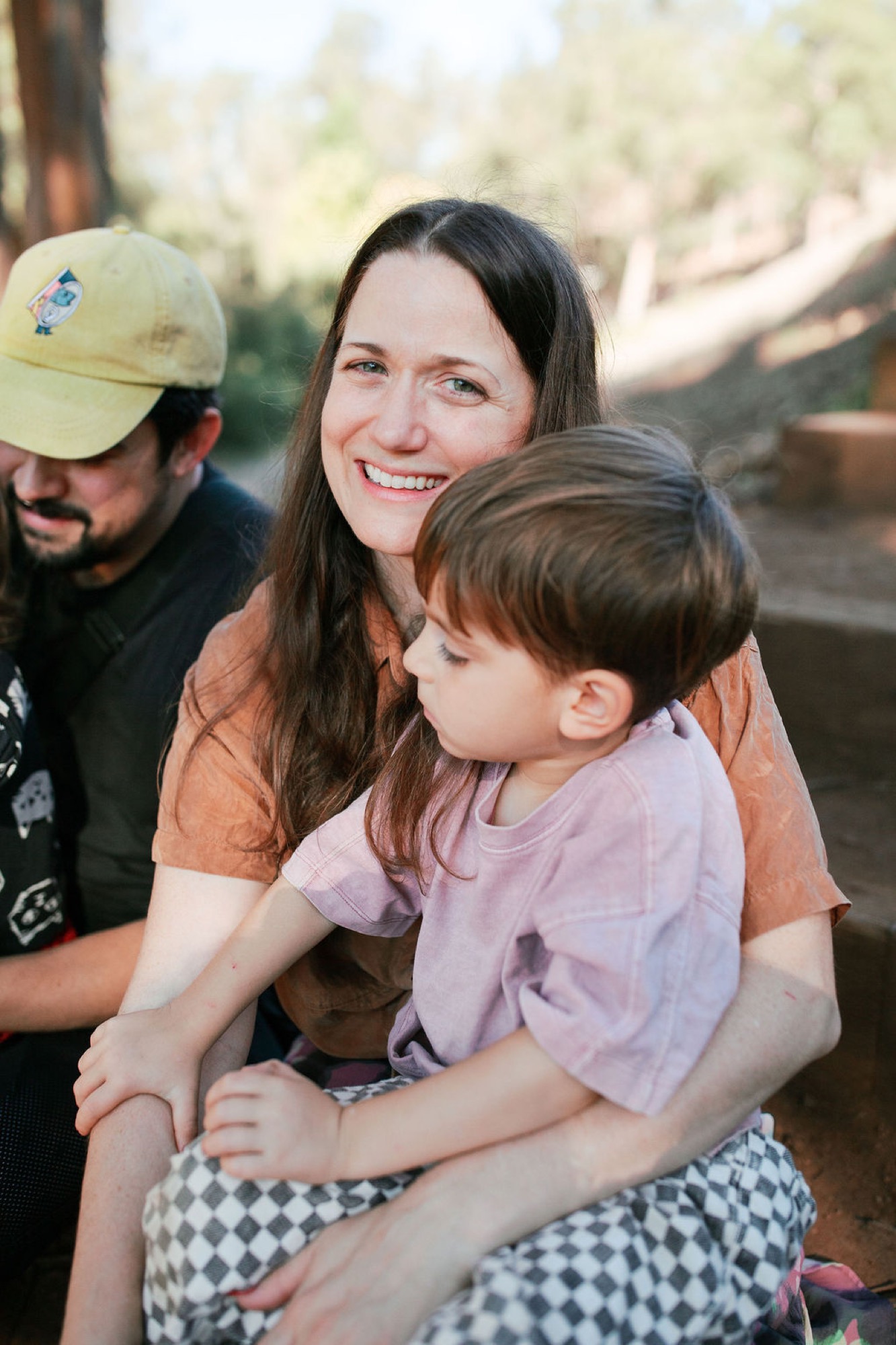 A woman in an orange shirt smiles warmly while embracing a young child in a purple shirt, with a bearded man in a yellow cap behind them in dappled forest light