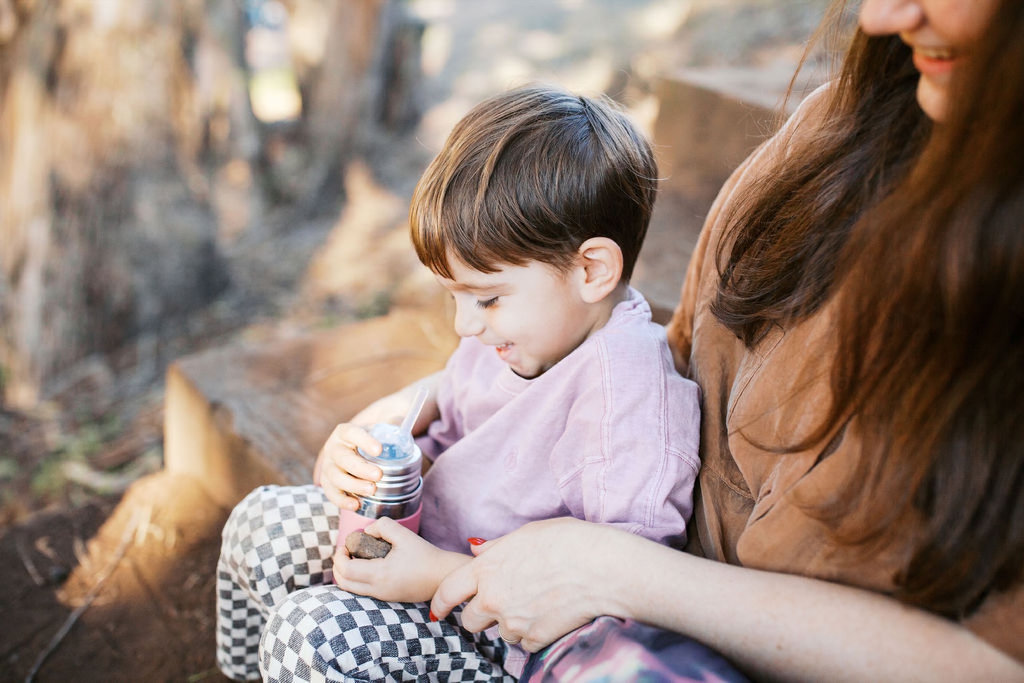 A young boy in a lavender shirt gazes downward while holding a pinecone, nestled against his mother's side on a sunlit hillside