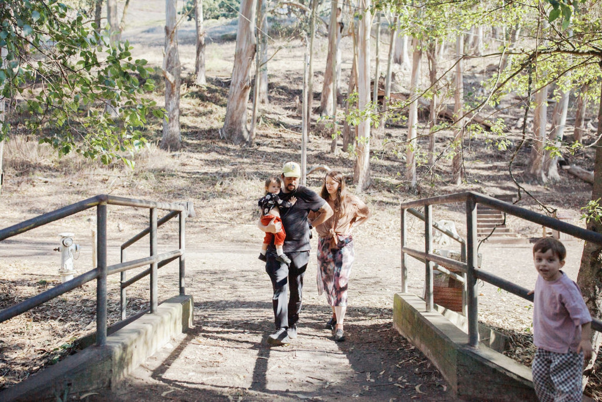 Family walks across a wooden bridge on a tree-lined trail, children in colorful clothing moving toward the camera with dappled forest light