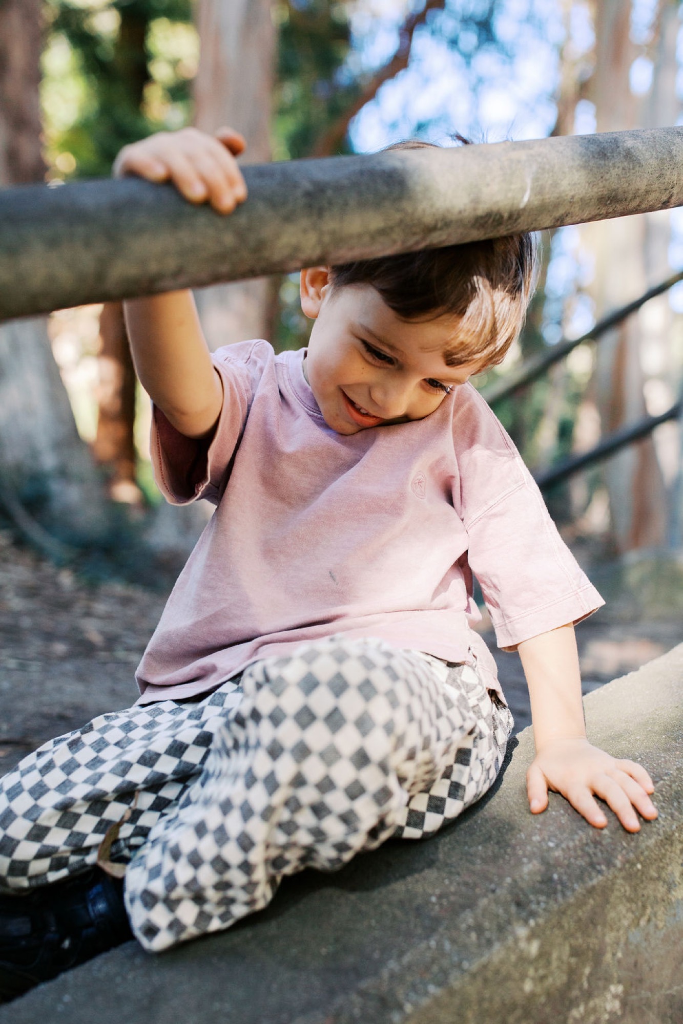 A young child in a lavender shirt hangs from a thick wooden beam, grinning with joy as dappled sunlight filters through the trees