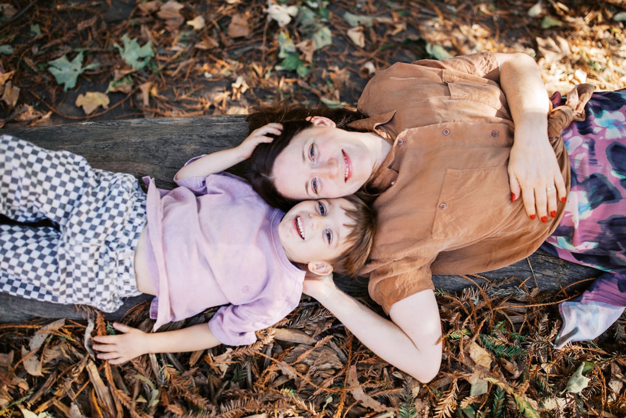 Two children lie on their backs on fallen leaves, gazing upward with gentle smiles as dappled sunlight filters through the forest canopy