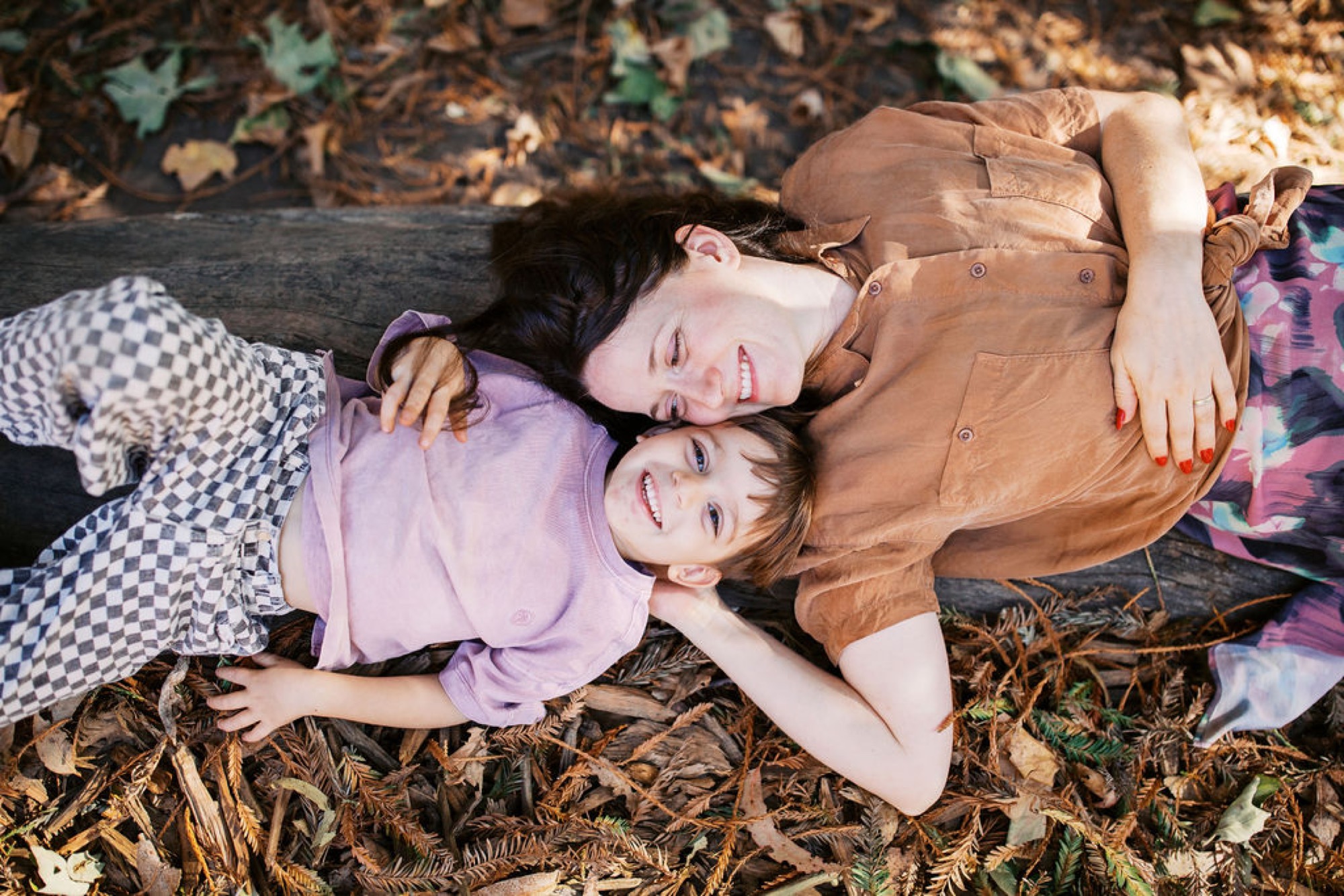 Two children lie peacefully on autumn leaf litter, one in lavender and one in tan, heads tilted toward each other in tender connection