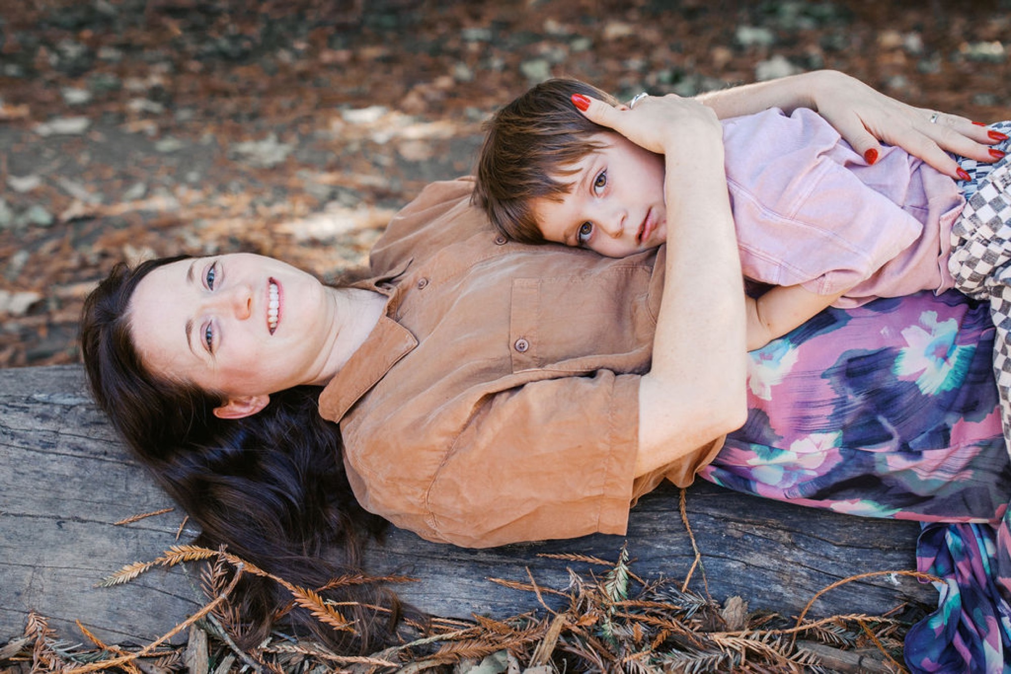 Mother and young child lying on forest ground, child resting on mother's chest with gentle expressions amid natural pine needles