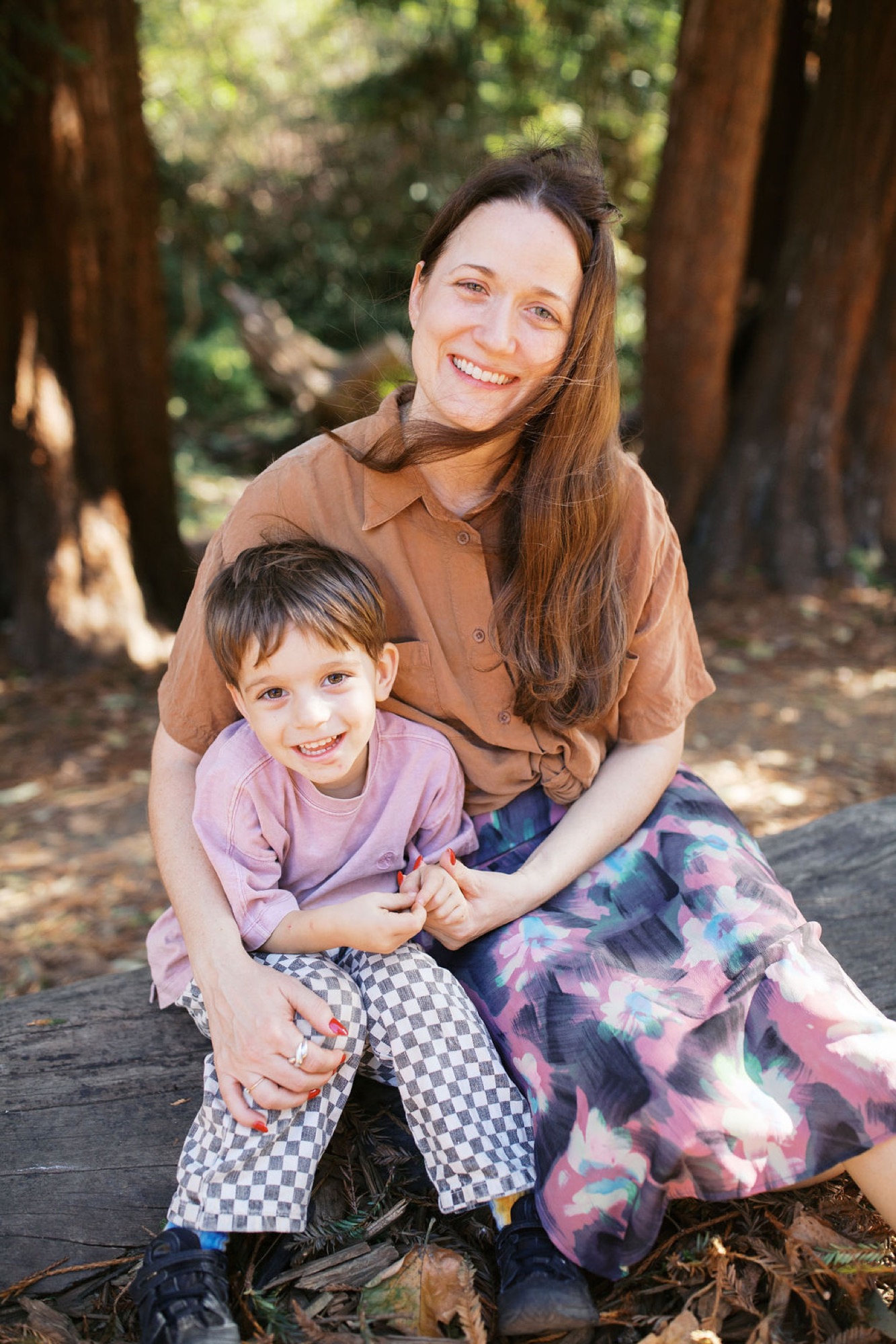 A woman and young boy smile warmly while sitting together on a log surrounded by towering redwoods dappled with golden sunlight