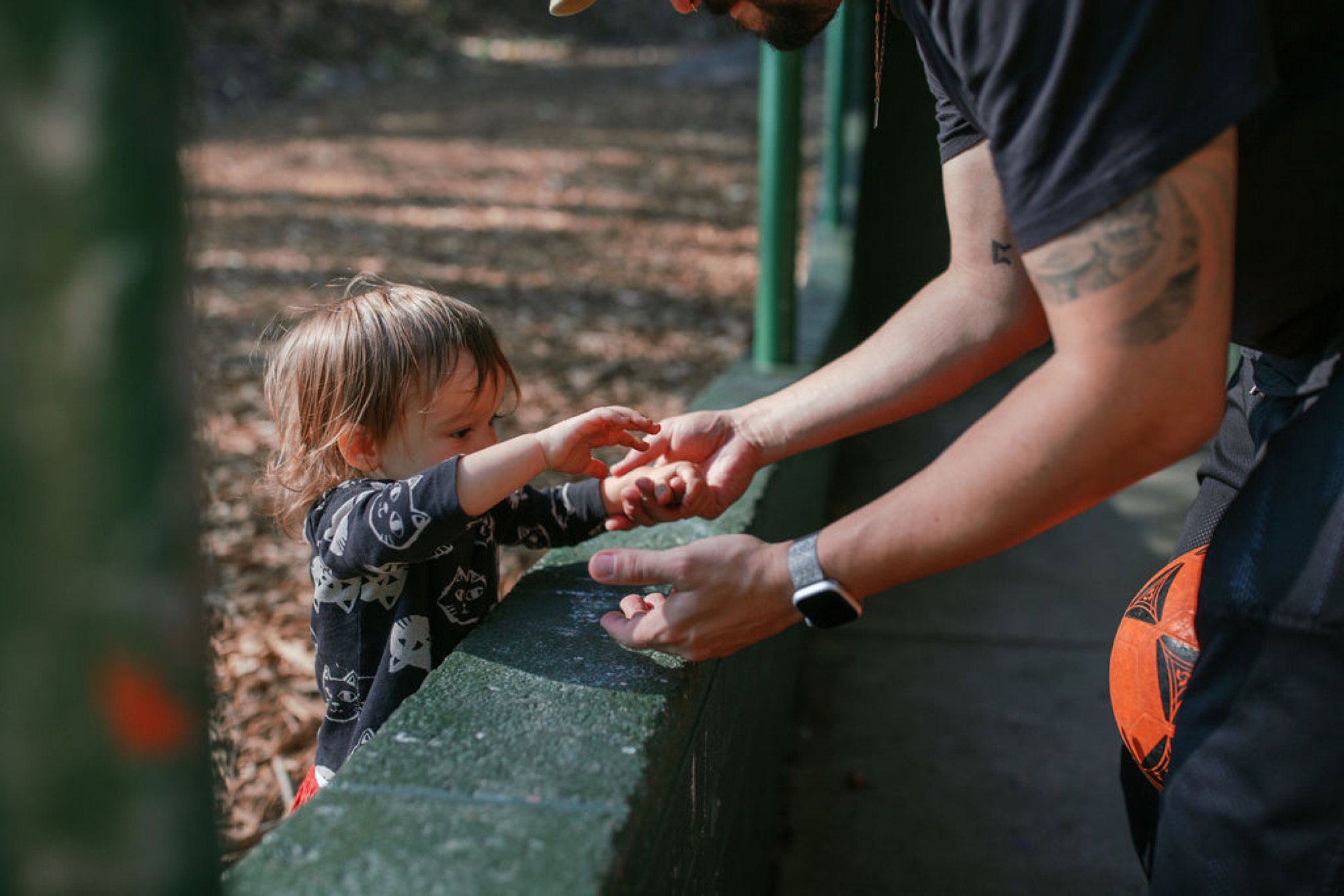A tattooed adult's hands guide a toddler's fingers along a weathered wooden rail in dappled sunlight