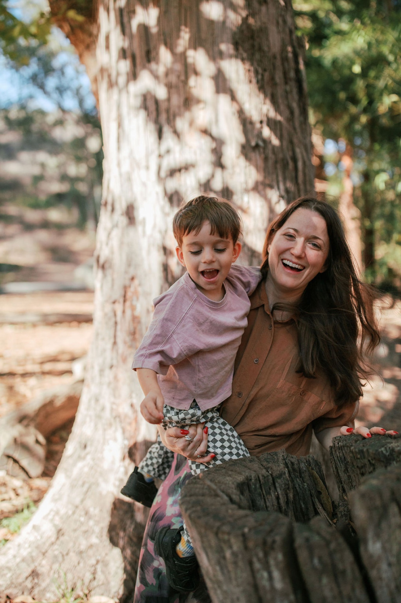 A woman and young child laugh together while sitting against a weathered eucalyptus tree, dappled sunlight casting shadows across their cheerful faces