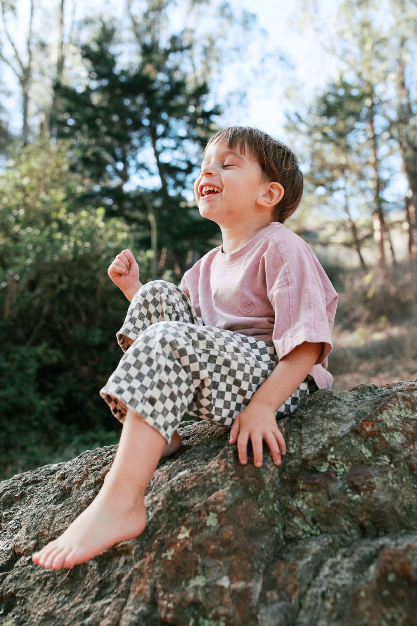 A laughing child in a pink shirt sits barefoot on a moss-covered boulder, gazing upward with pure joy as dappled sunlight filters through the forest canopy