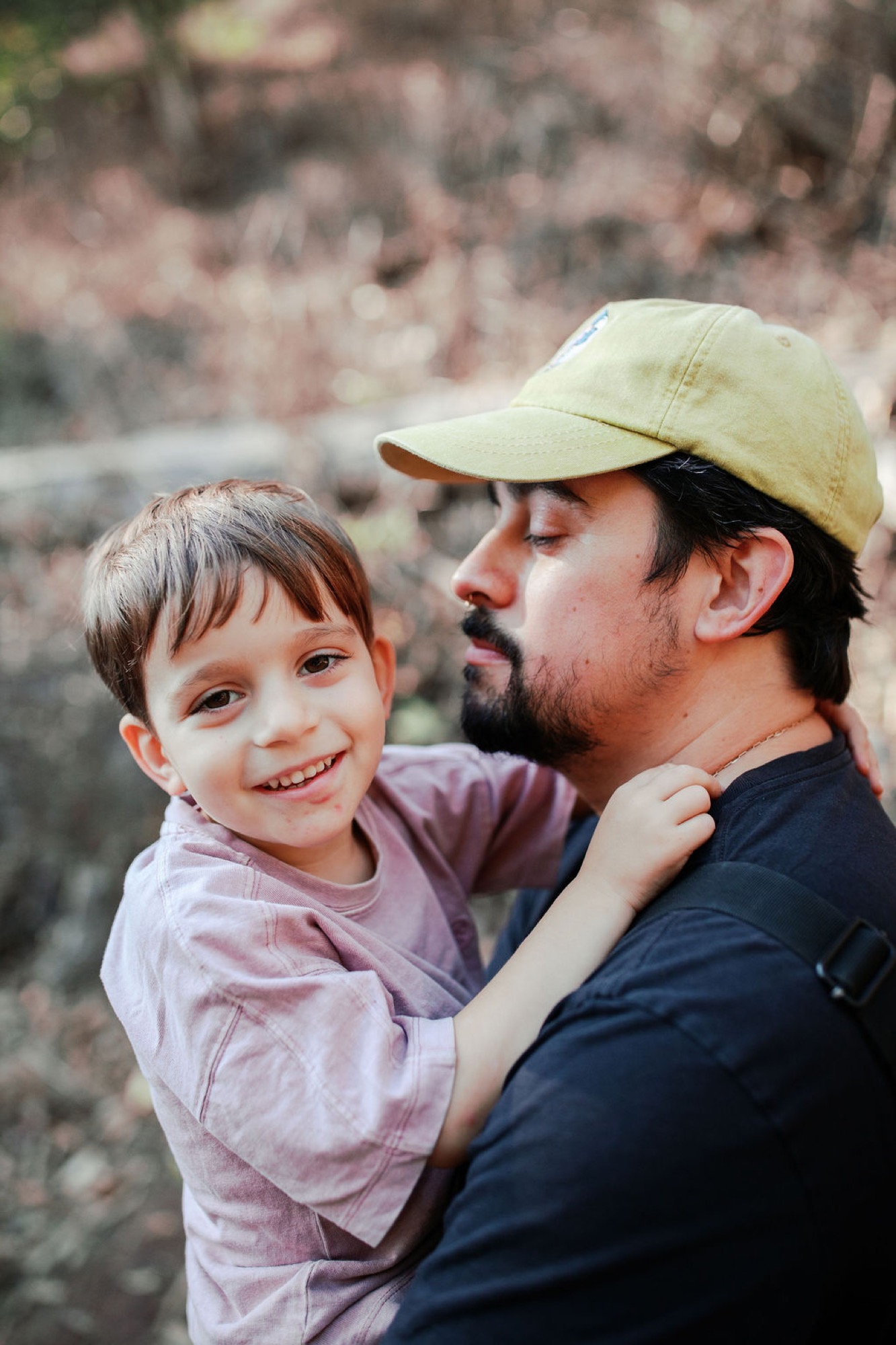 A bearded man in a yellow cap holds a smiling toddler against a blurred rocky backdrop, both gazing downward in a moment of quiet connection