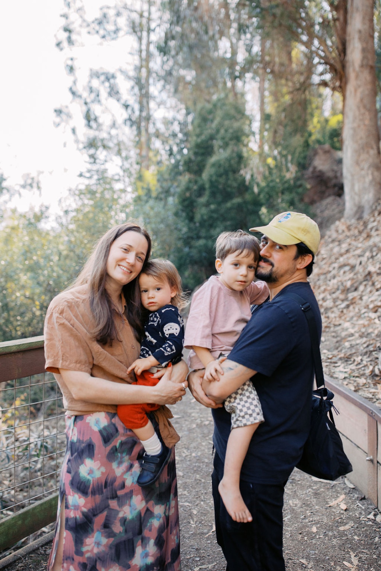 A family of four stands together on a forested trail, with warm afternoon light filtering through tall trees behind them