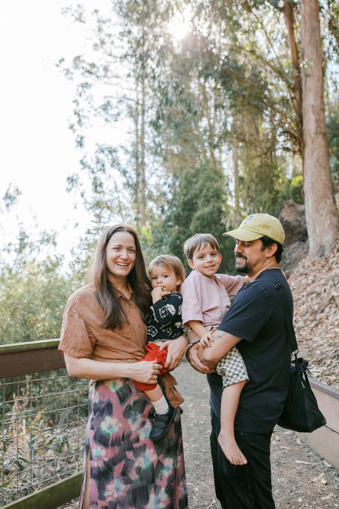 A smiling family of four stands together on a wooded trail, with tall eucalyptus trees backlit by bright sunlight filtering through the canopy