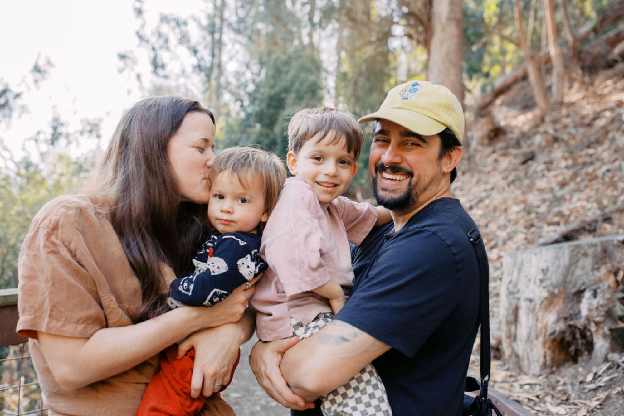 A smiling family of four nestles together on a wooded trail, dappled sunlight filtering through tall trees behind them