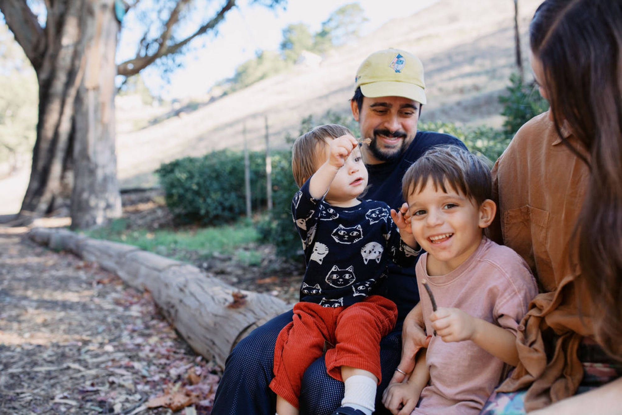 A bearded father in a cream cap holds his young daughter while his son leans in close, all smiling beneath dappled tree shade with hillside vineyards visible behind them