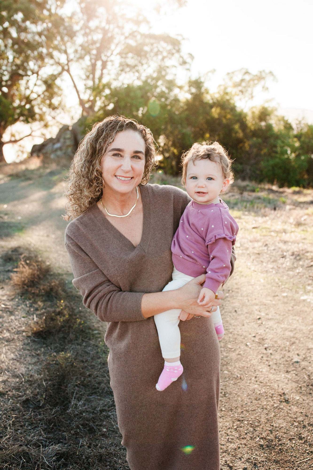 A woman in a taupe cardigan holds her toddler in a purple top along a sunlit dirt path, with tall trees backlighting the warm golden hour