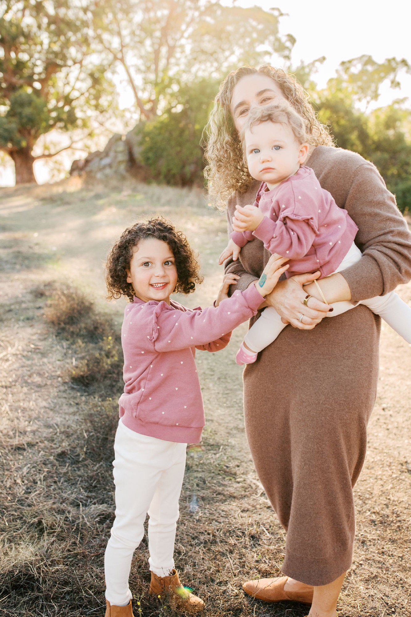 A mother playfully holds her toddler while her older daughter smiles beside them on a sunlit dirt path lined with golden trees
