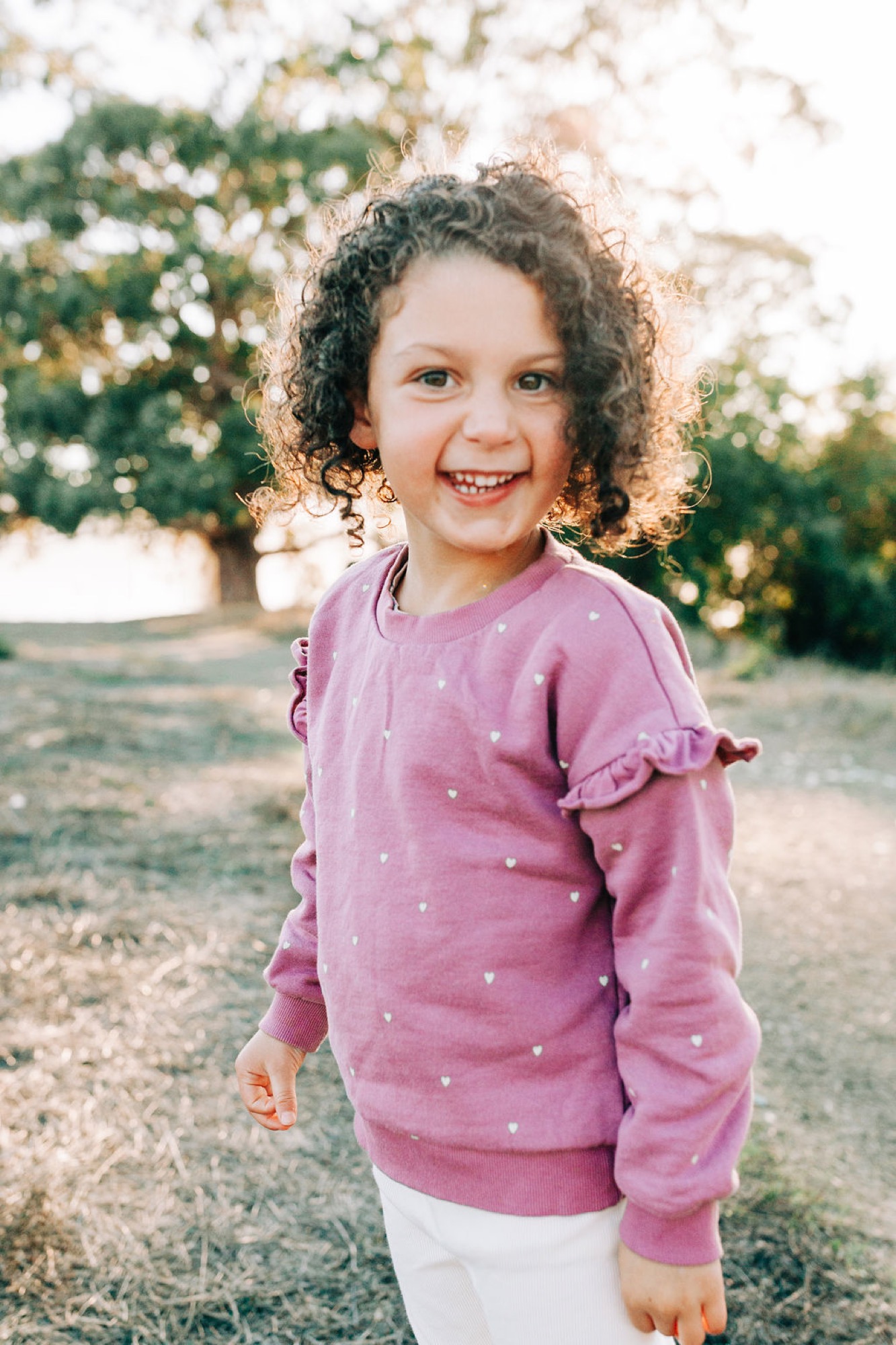 Young girl with curly hair beams at the camera in a pink heart-patterned sweater against blooming white flowers and green foliage