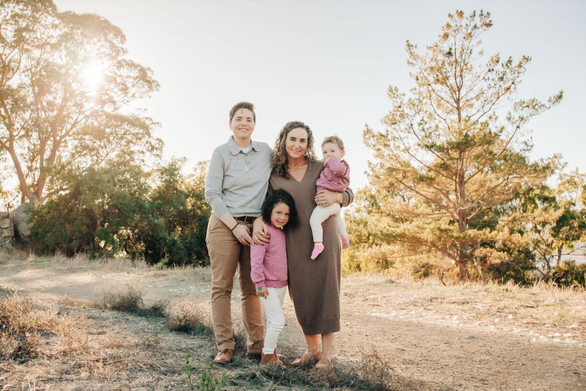 A family of four stands on a dusty trail surrounded by golden autumn trees, the mother smiling warmly while holding her youngest child in pink