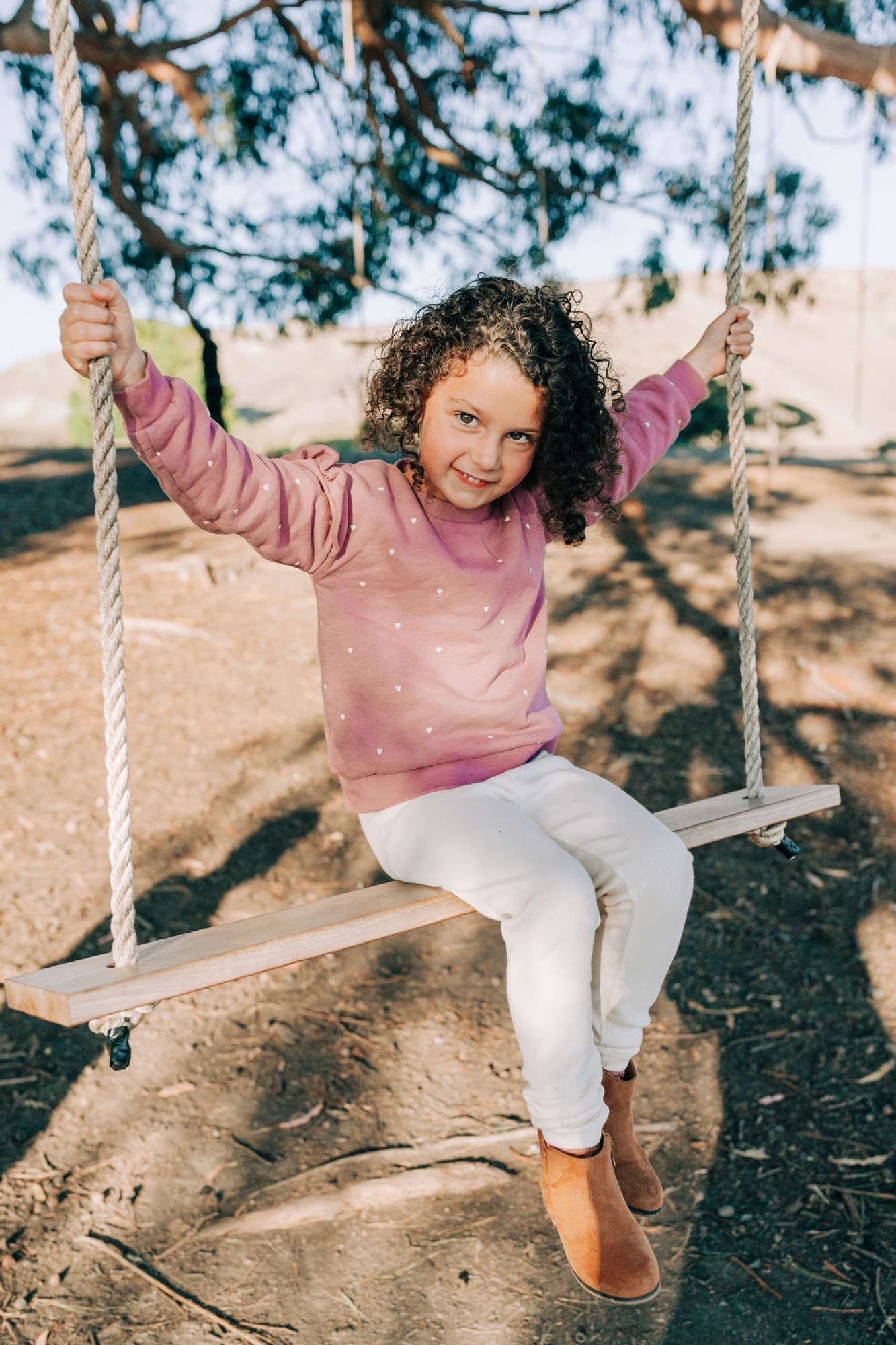 A curly-haired child grins widely while swinging from a rope beneath oak tree dappled shade