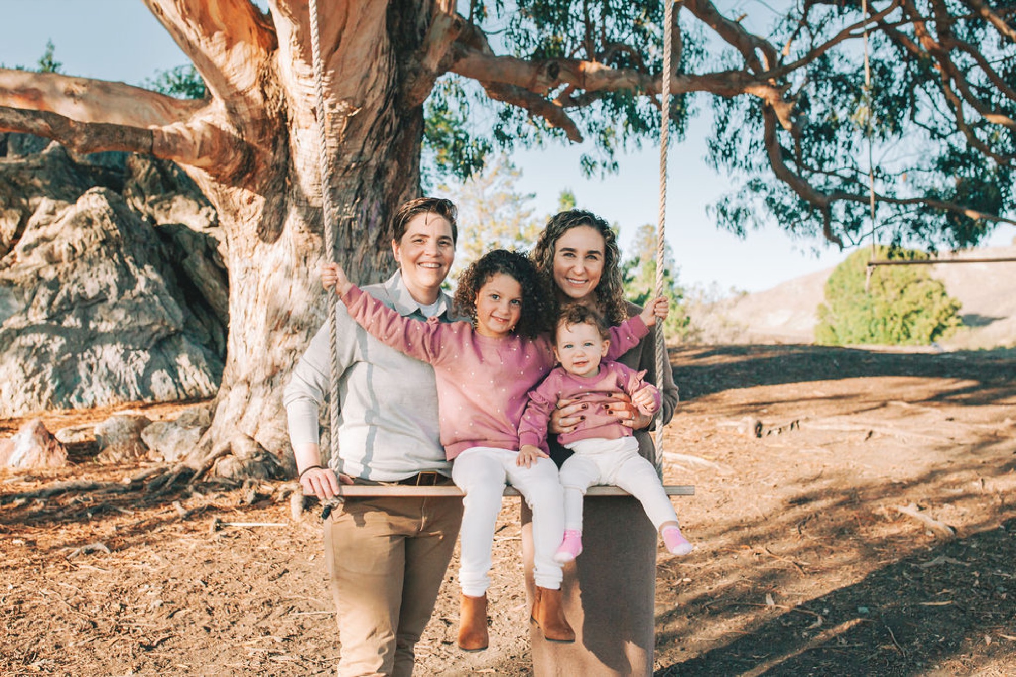A family of four smiles warmly on a rope swing beneath a massive eucalyptus tree, bathed in golden afternoon light with rolling hills behind