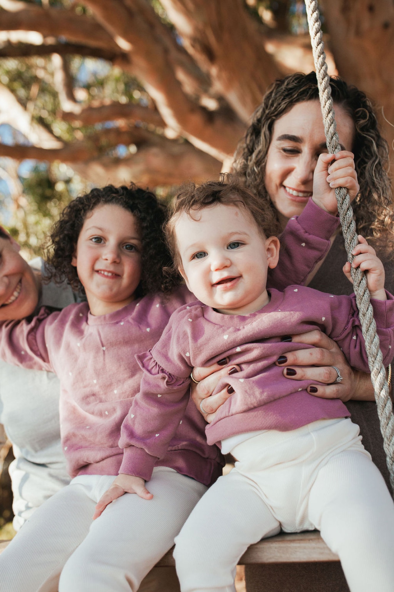 Three children in coordinated pink tops grip a rope swing, beaming with genuine joy against rustic wooden beams and dappled sunlight