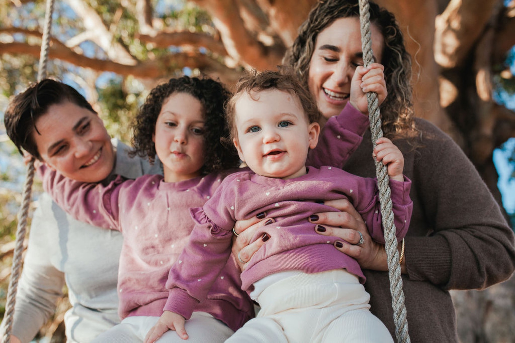 Four females on a rope swing smile warmly in dappled sunlight beneath sprawling oak branches, the youngest child gleaming with joy